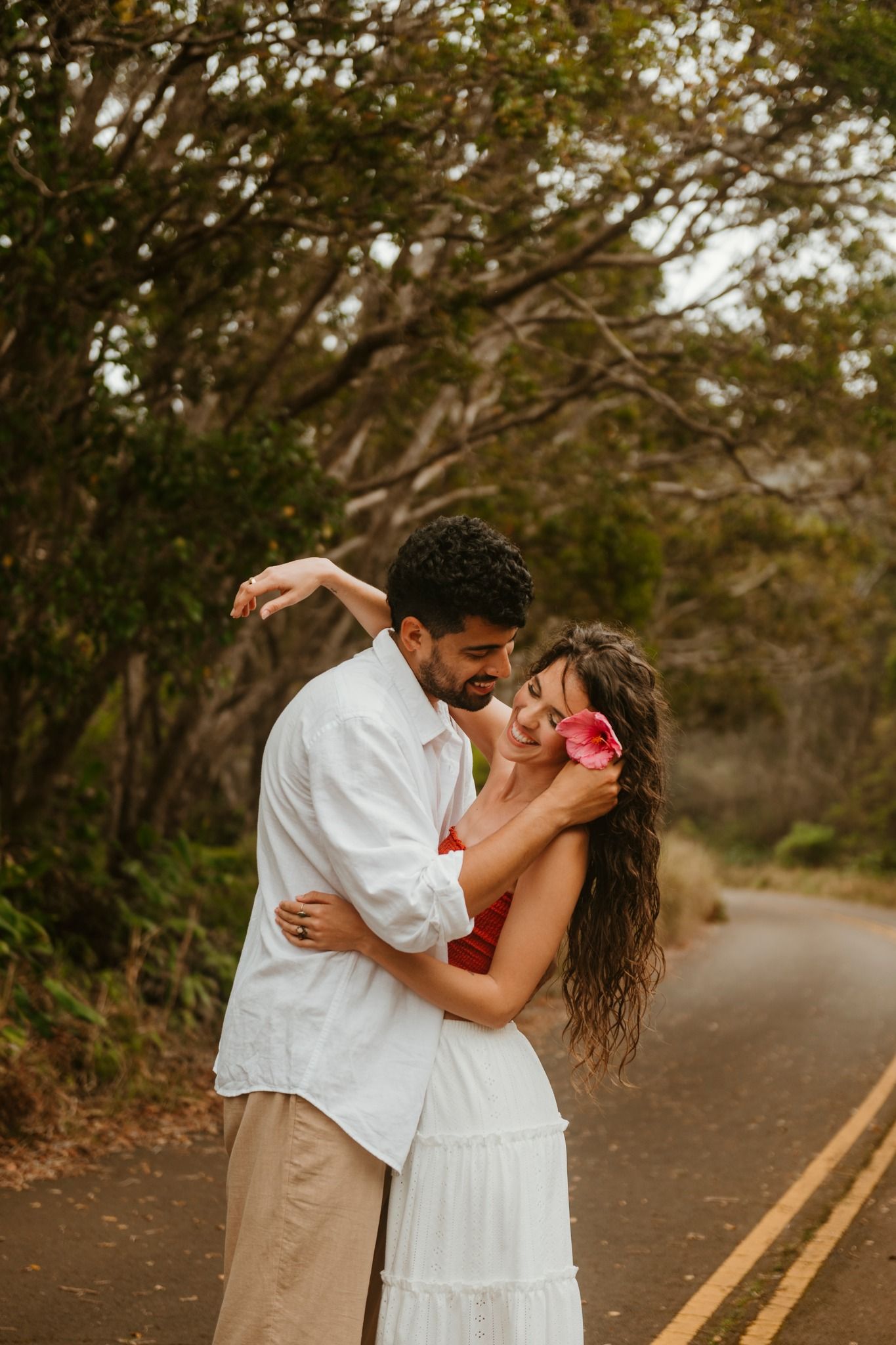 Romantic couple embracing on a scenic road surrounded by lush trees, woman holding a pink flower in her hair.