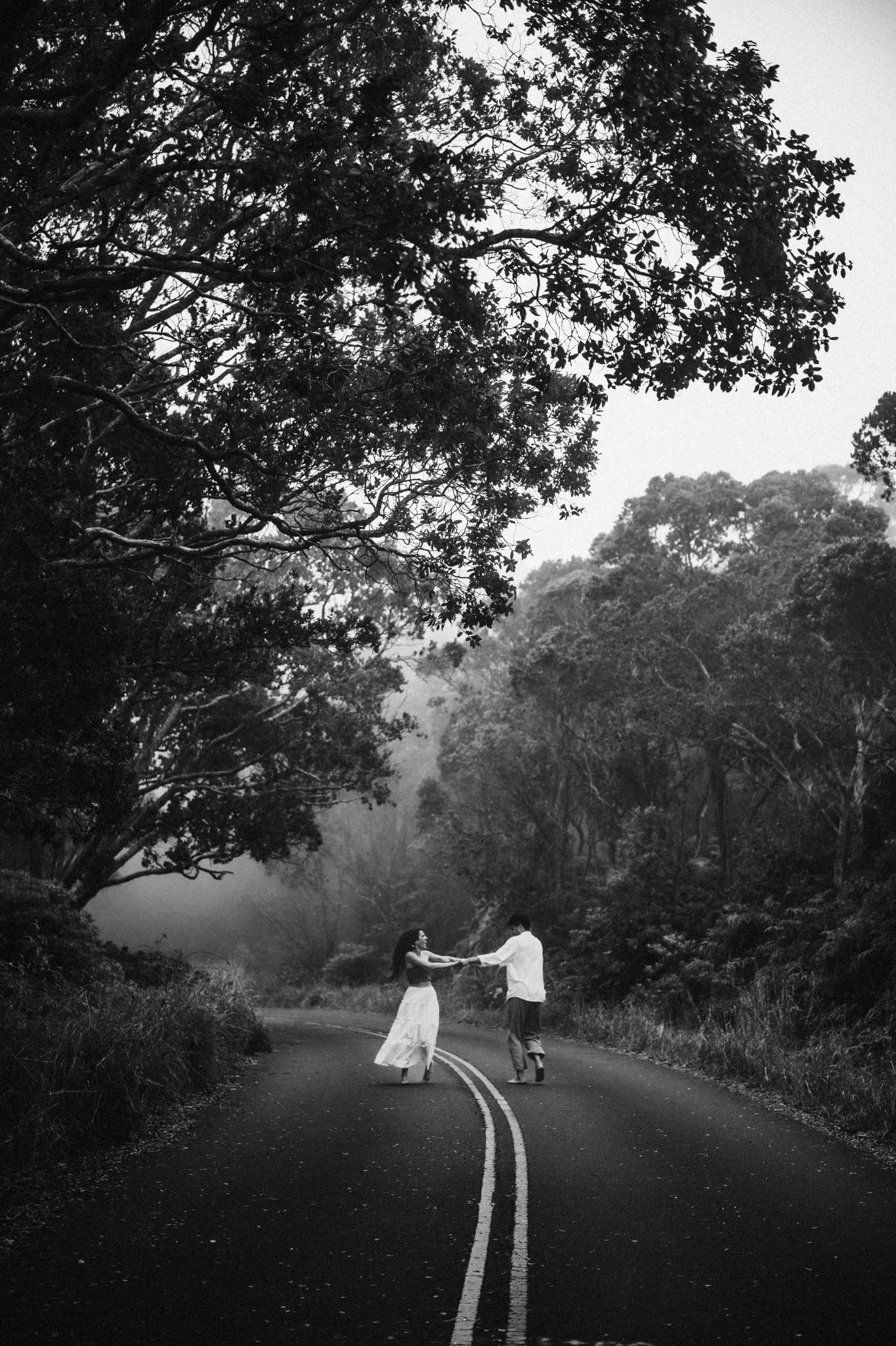 Black and white photo of a couple dancing on a misty tree-lined road.