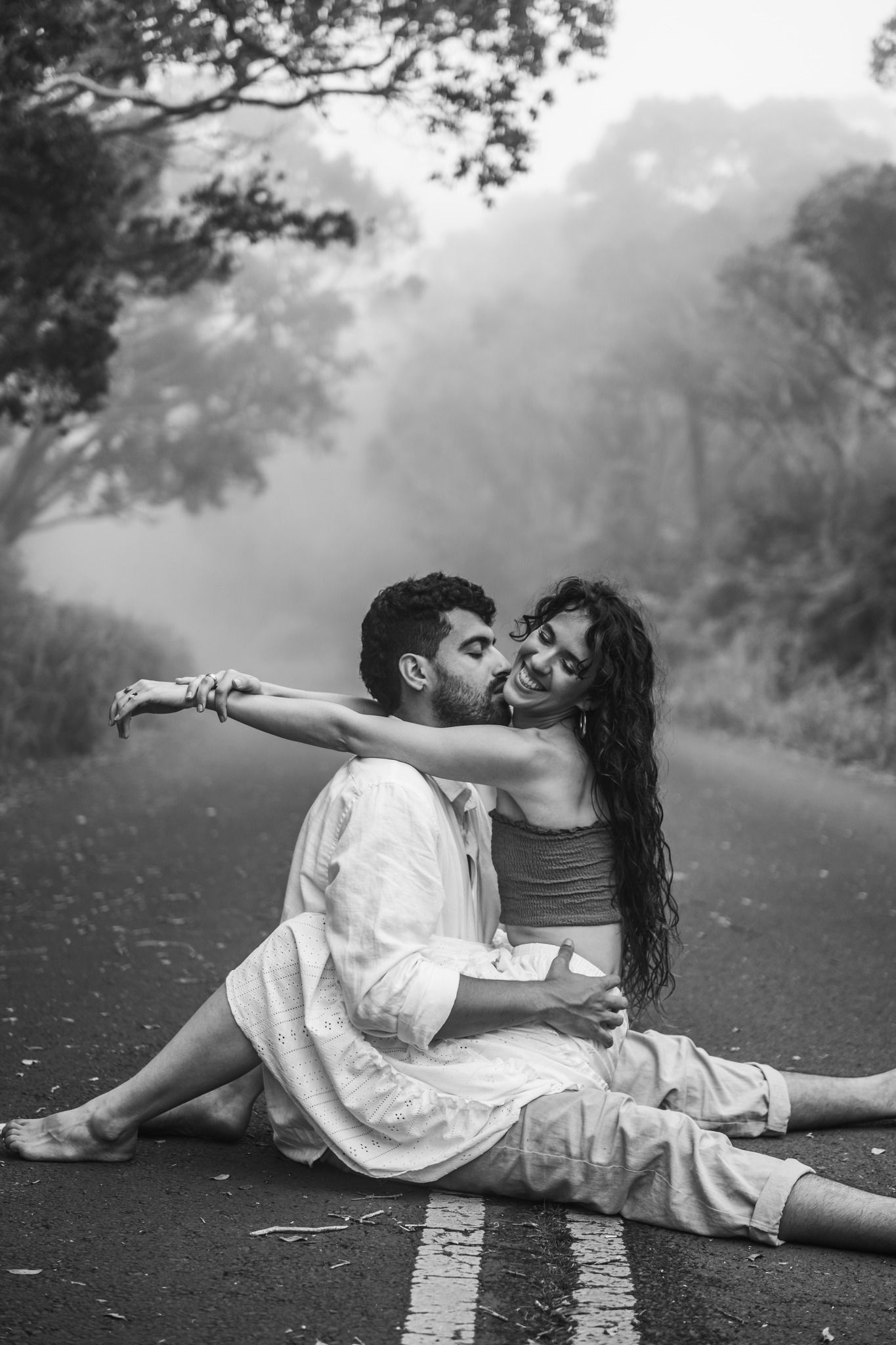 Couple sitting on road centerline in black and white, sharing a romantic moment in fog.