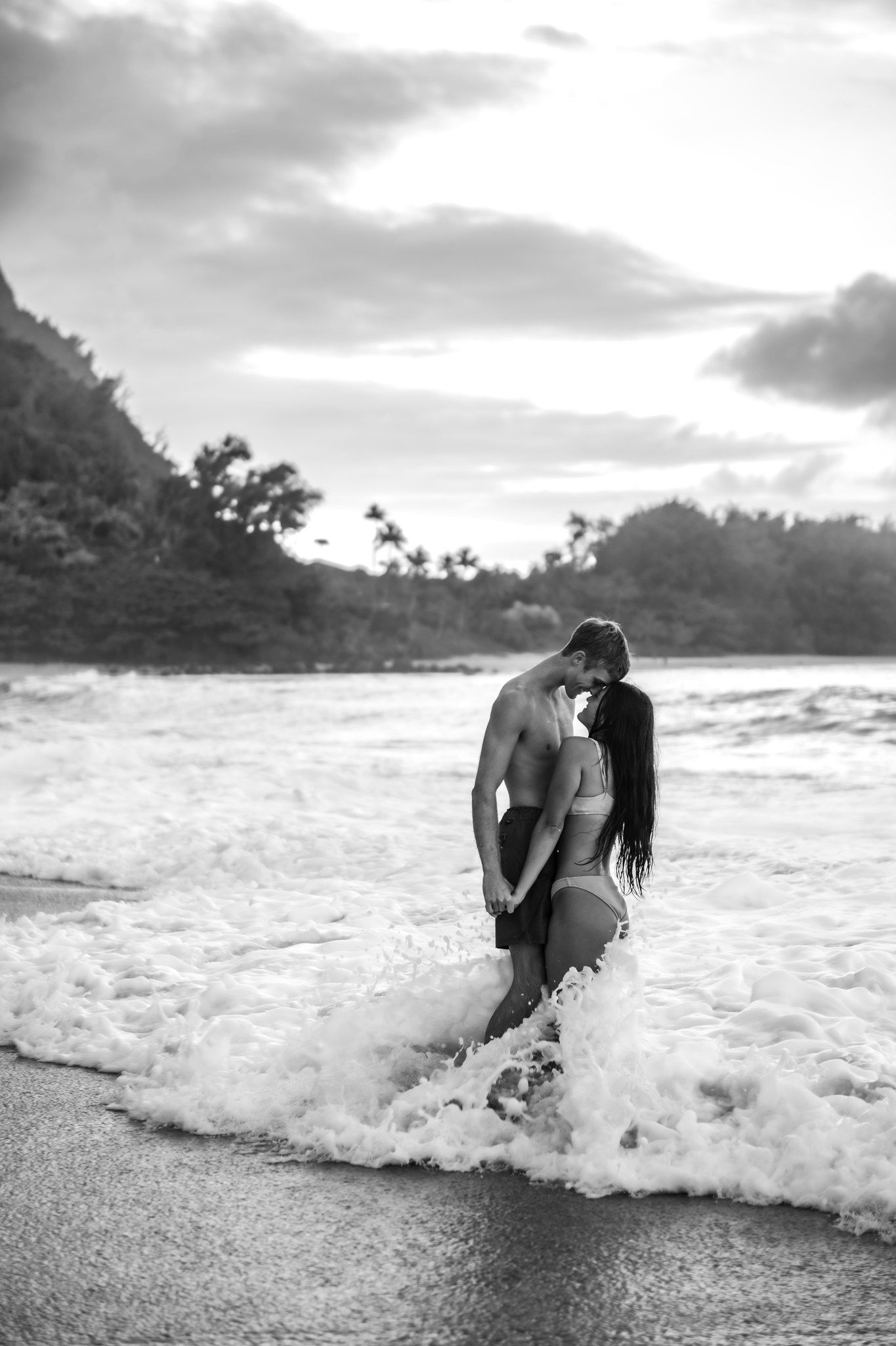 Black and white photo of a couple kissing in ocean waves at sunset on a tropical beach.