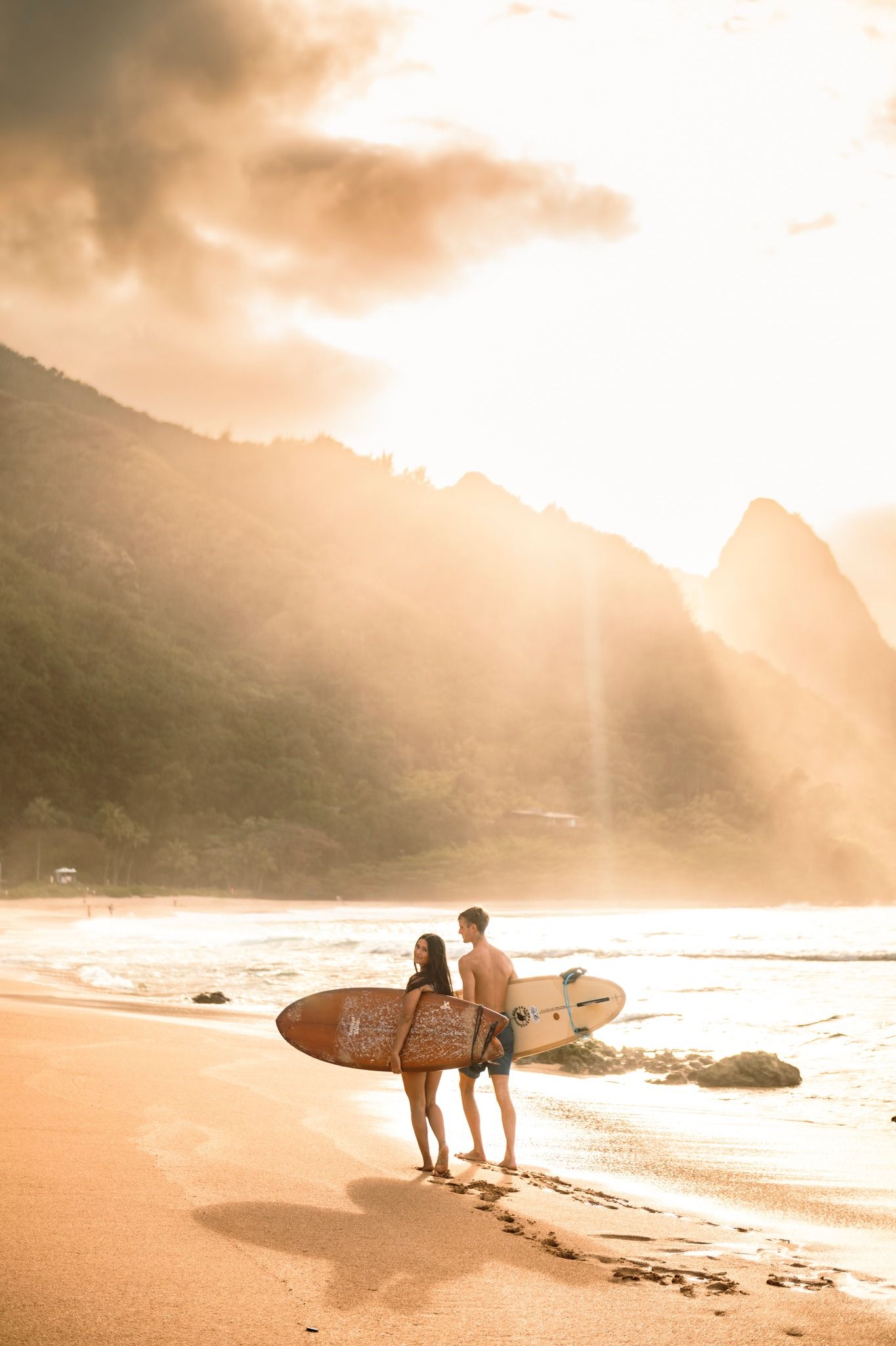 Couple holding surfboards on a golden sunlit beach with dramatic mountain backdrop.