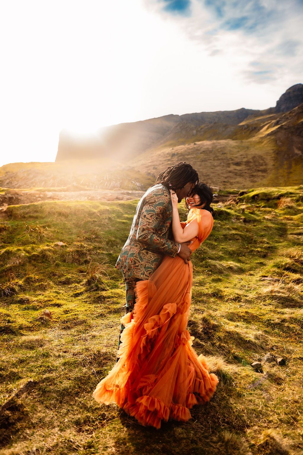 Couple embracing in an orange gown and patterned suit on a sunlit green hillside with misty mountains.
