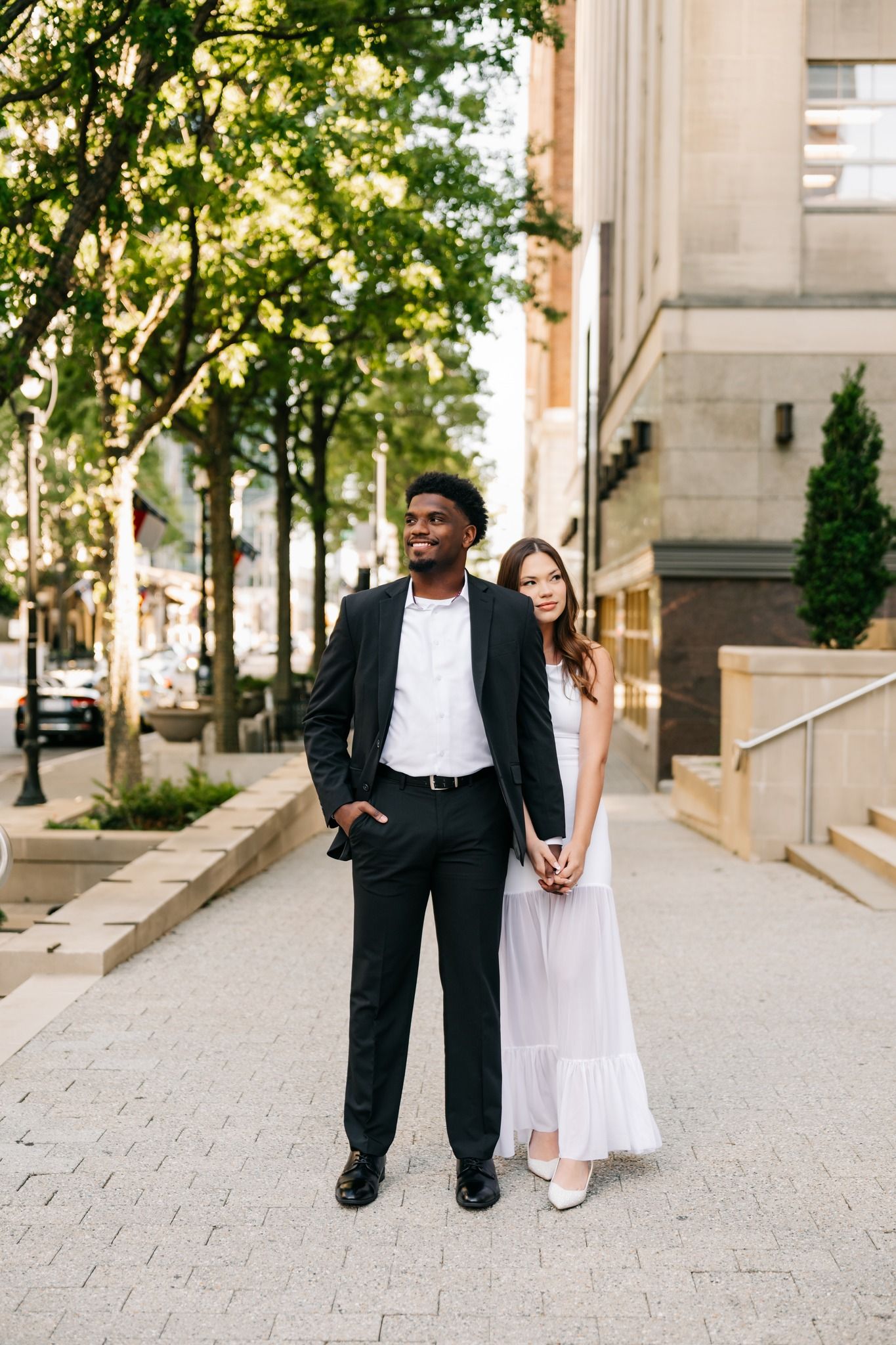 Interracial couple in formal attire holding hands on a city sidewalk, man in black suit, woman in white dress.