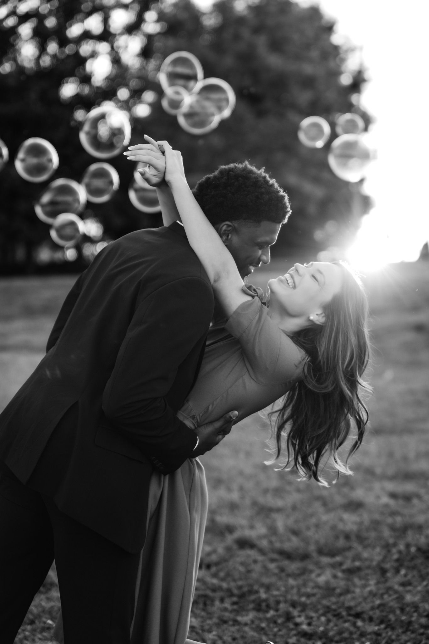 Black and white photo of a couple dancing and kissing outdoors surrounded by bubbles with sunlight in background.