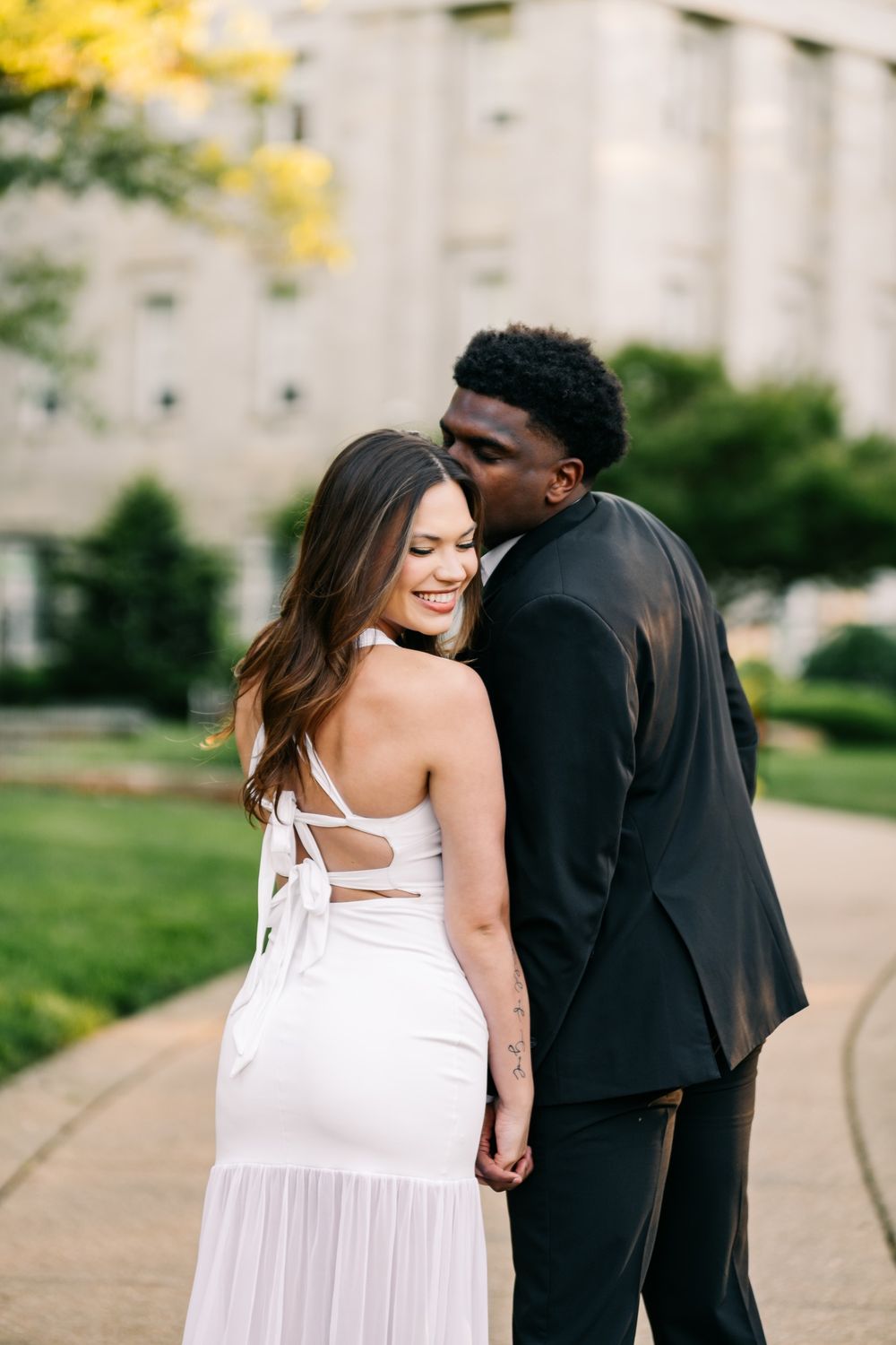 Couple in formal wear holding hands outdoors, woman in white dress smiling as man in black suit leans in close.