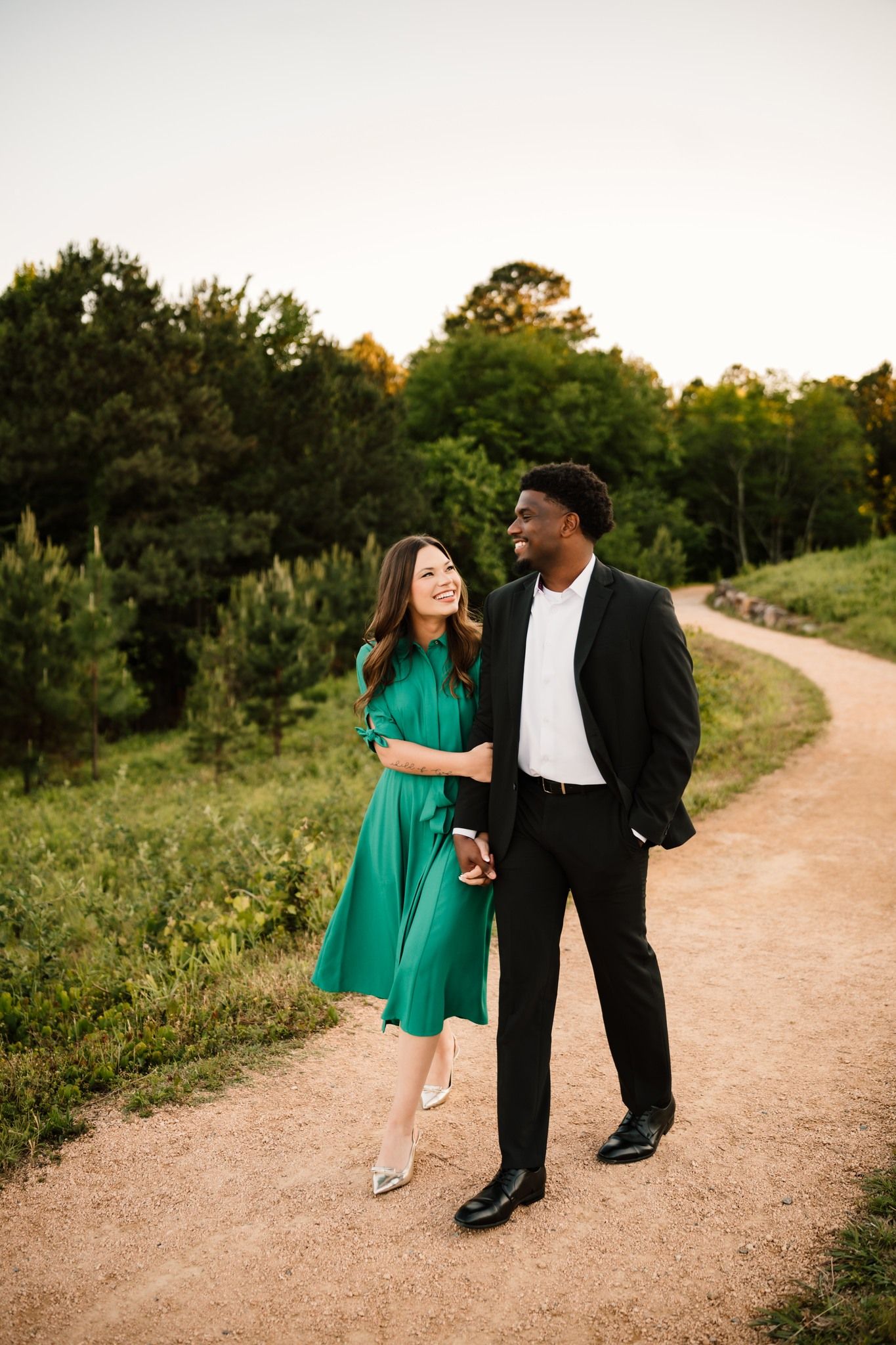 A couple walks hand-in-hand on a dirt path, woman in green dress and man in black suit, surrounded by lush green trees.
