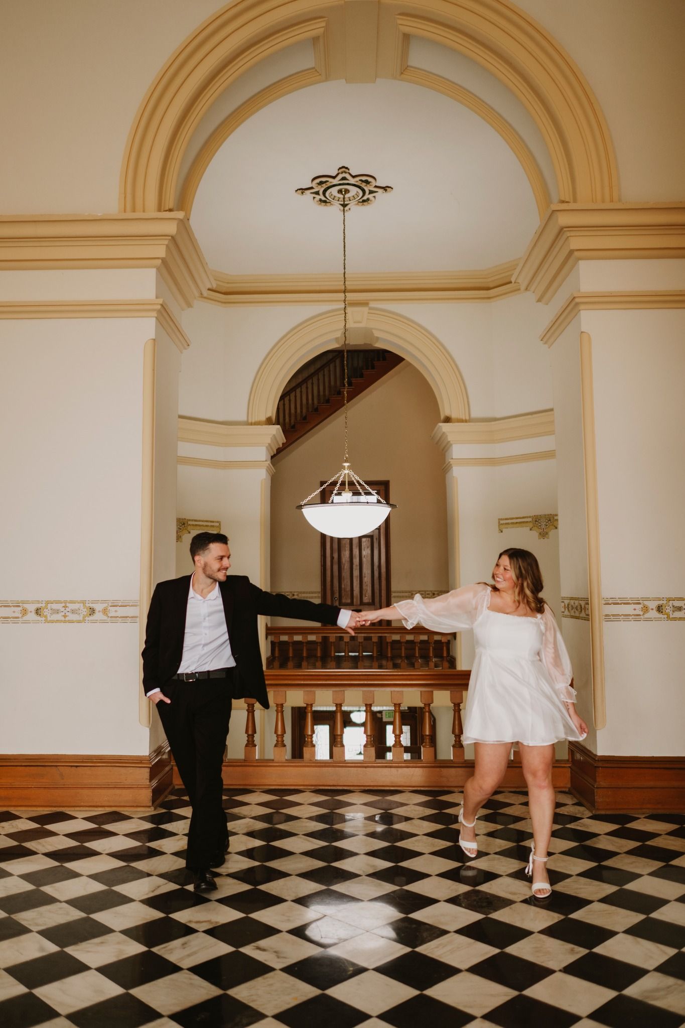 Couple dancing joyfully in elegant venue with checkered floors and ornate arched ceilings in white and black attire.