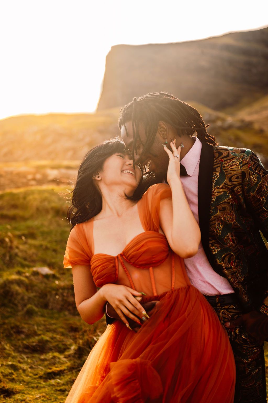 Couple embracing at golden hour, woman in orange tulle gown, man in patterned blazer, scenic highland cliffs backdrop.