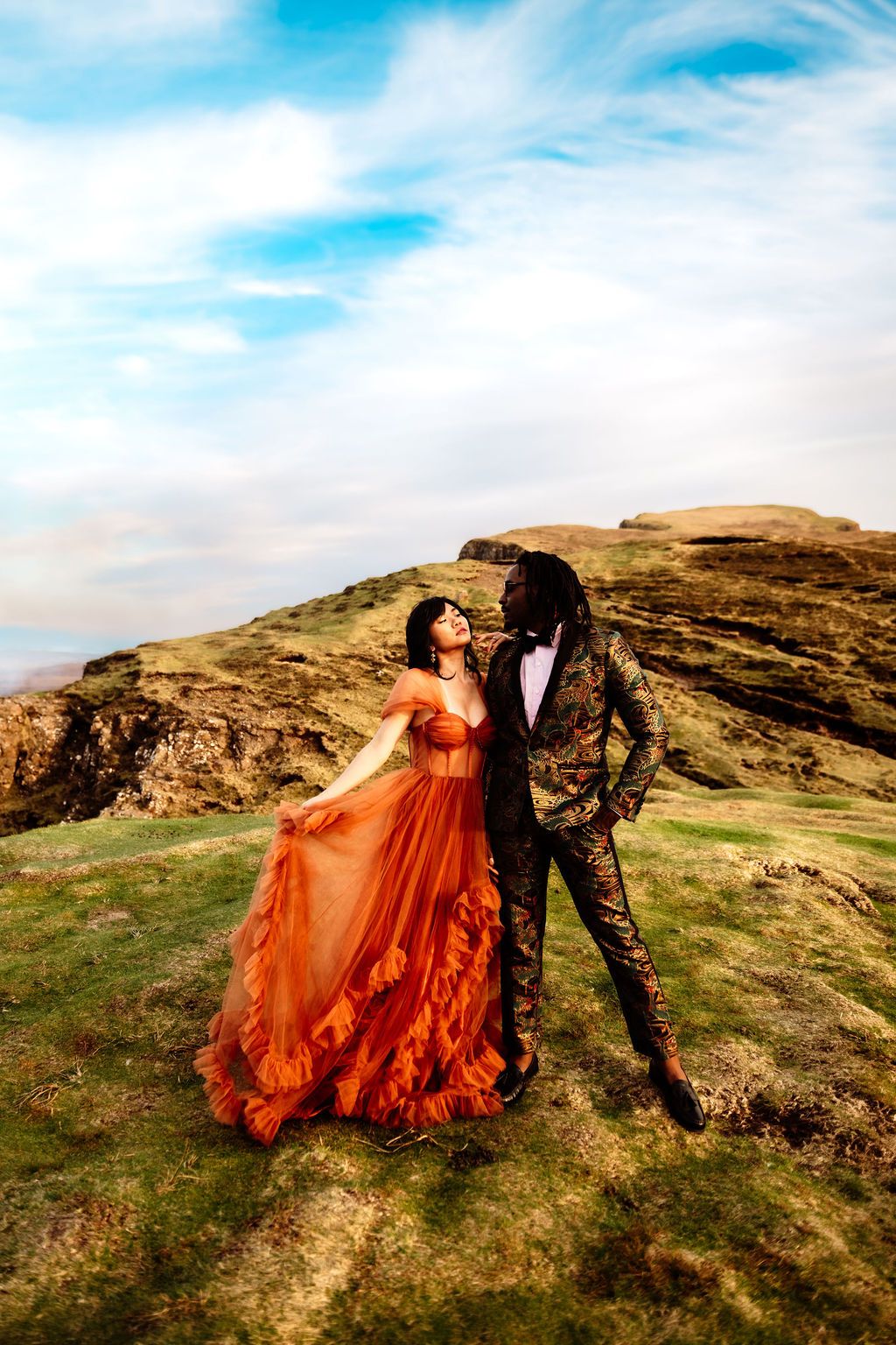 Couple in elegant attire poses on a scenic hilltop, woman in flowing orange gown, man in patterned suit, blue sky backdrop.