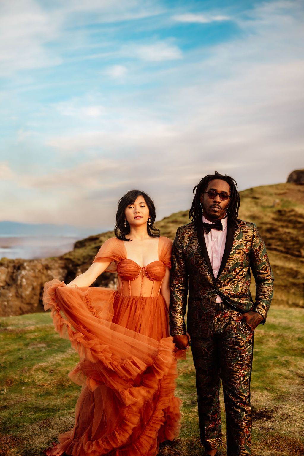 Couple in elegant attire poses outdoors, woman in rust tulle gown, man in floral suit, scenic coastal backdrop.
