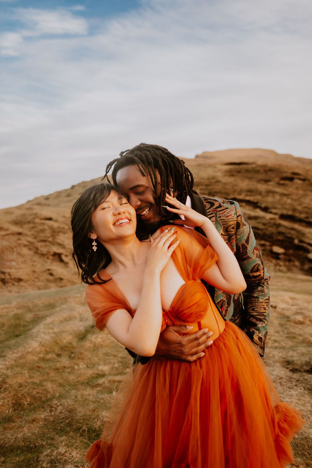 Joyful interracial couple embracing outdoors, woman in orange tulle gown laughing as man with dreadlocks kisses her cheek.