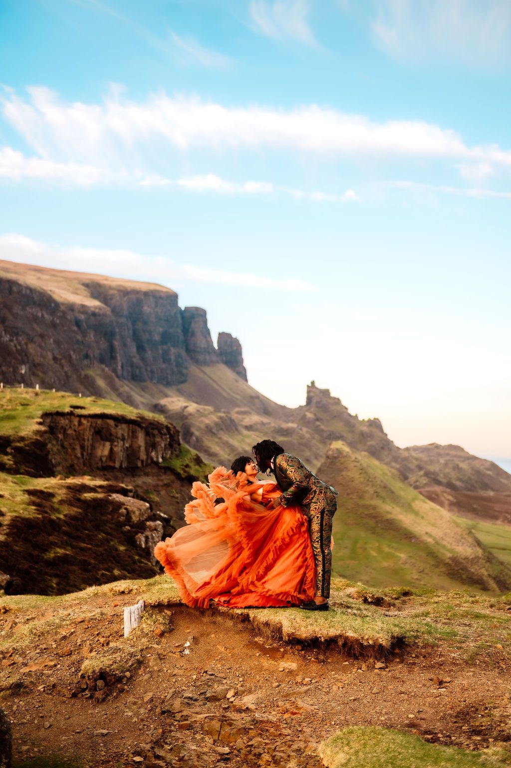 Couple in vibrant orange gown and ornate jacket sitting on rocky highland cliffs with dramatic mountain landscape behind them.