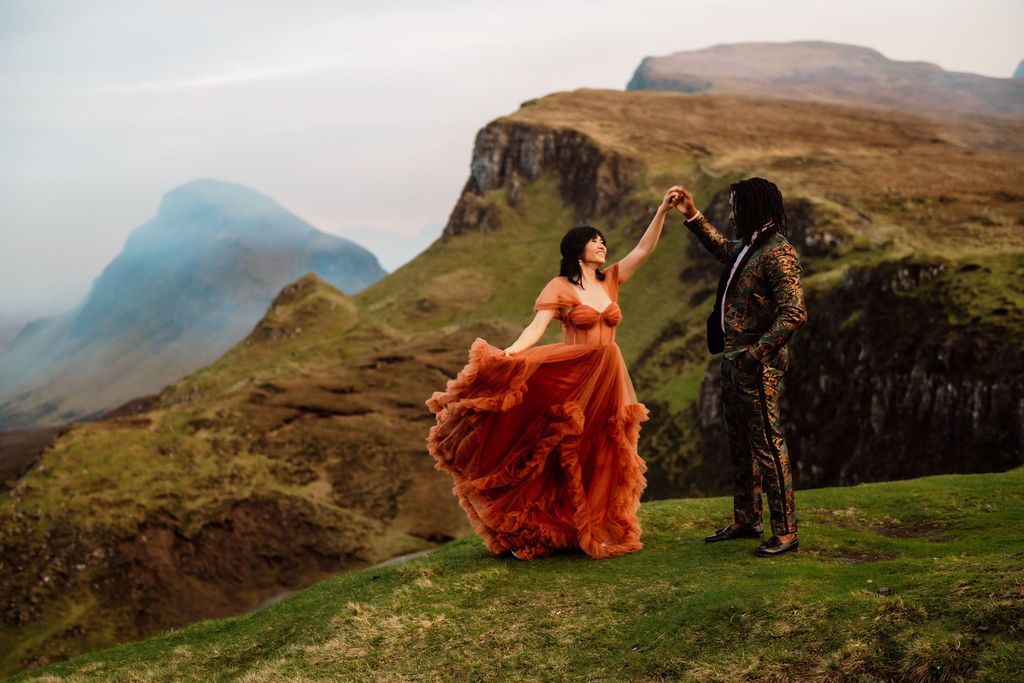 Couple dancing on Scottish Highland cliffs, woman in flowing orange gown, misty mountains backdrop.