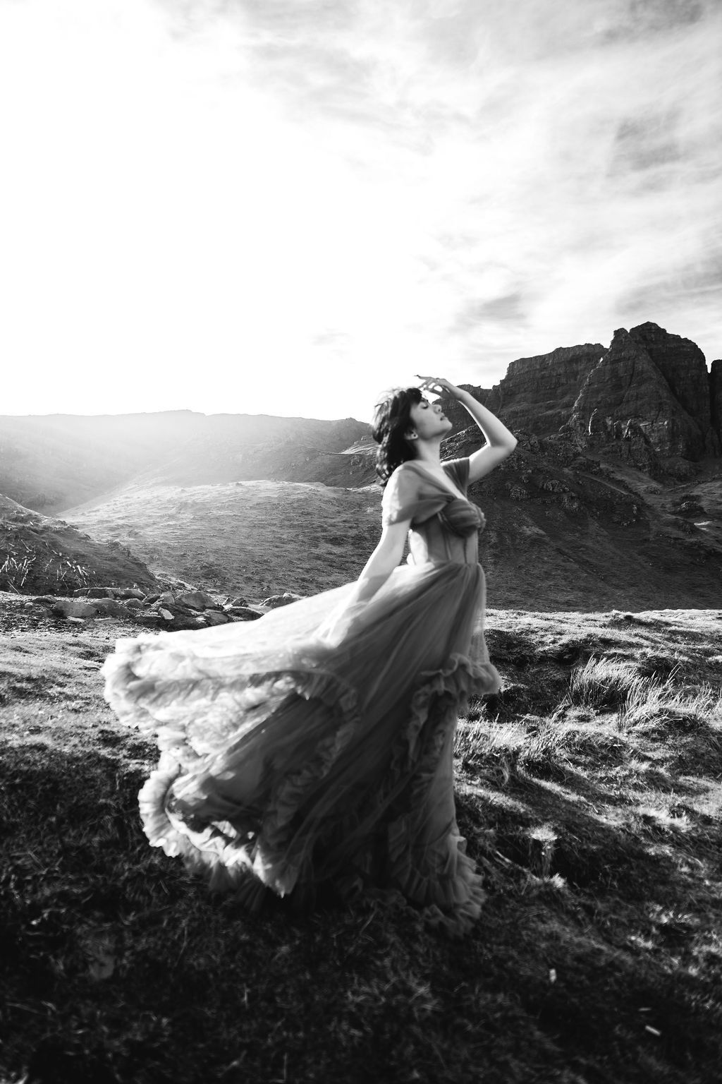 Woman in flowing gown standing on rocky highland landscape in dramatic black and white photo.