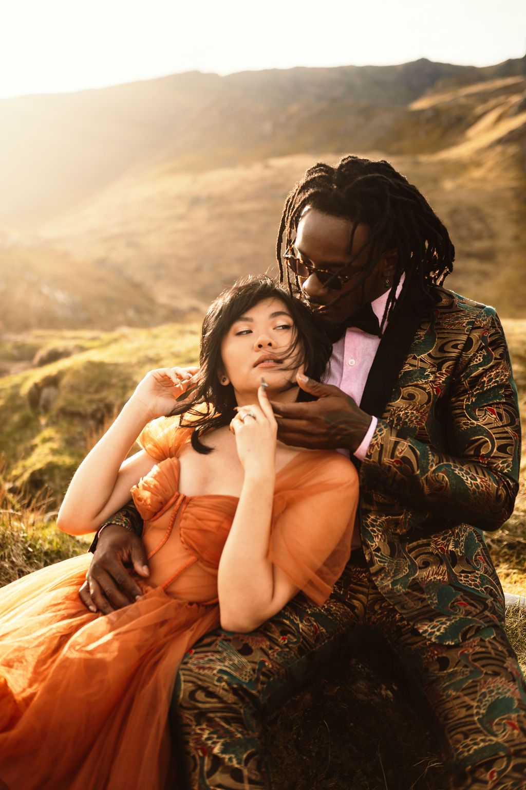 Interracial couple in elegant attire sitting on a hillside at golden hour, woman in orange gown, man in patterned suit.