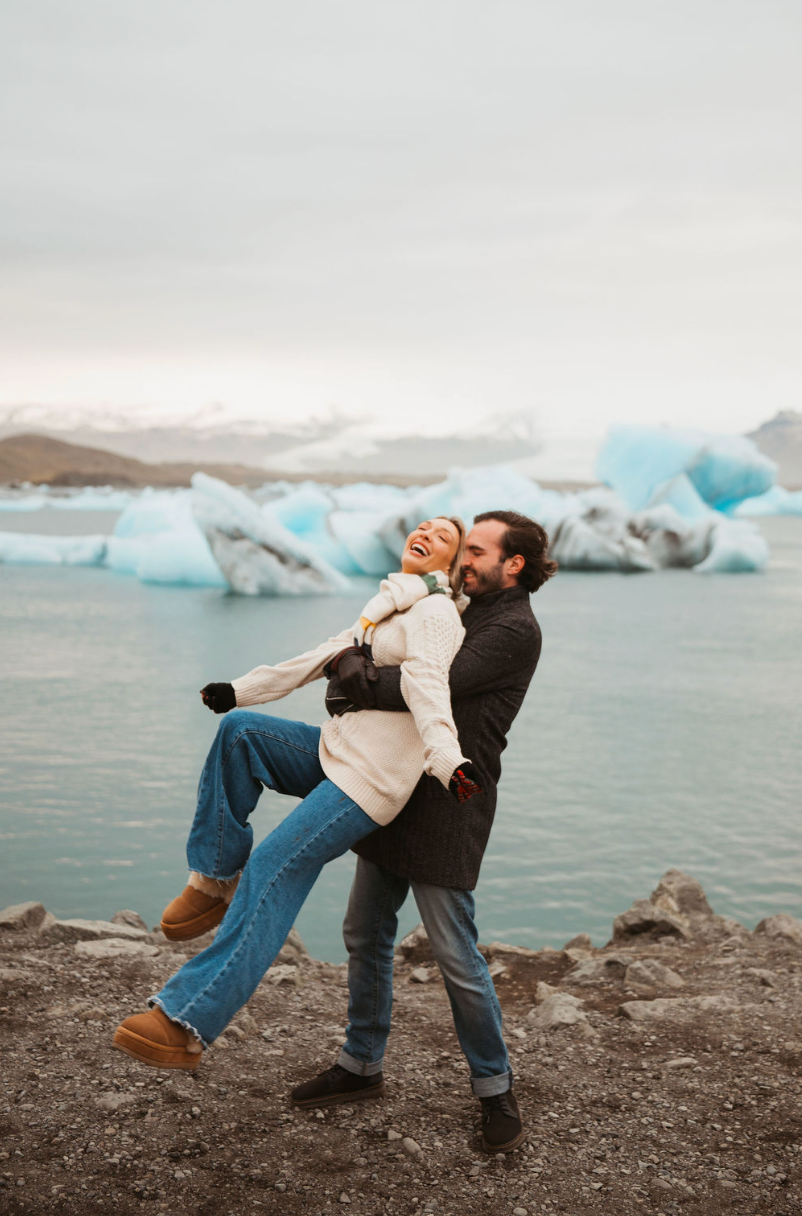 A joyful couple embraces near a glacial lagoon with blue icebergs in Iceland, as the man lifts the laughing woman.