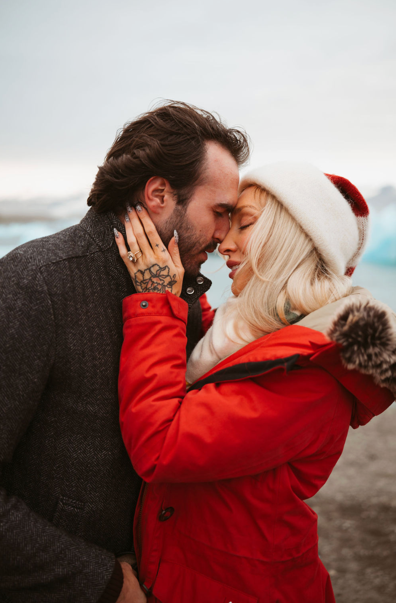 A couple shares a romantic moment outdoors, woman in red coat and Santa hat, man in gray wool coat, winter backdrop.