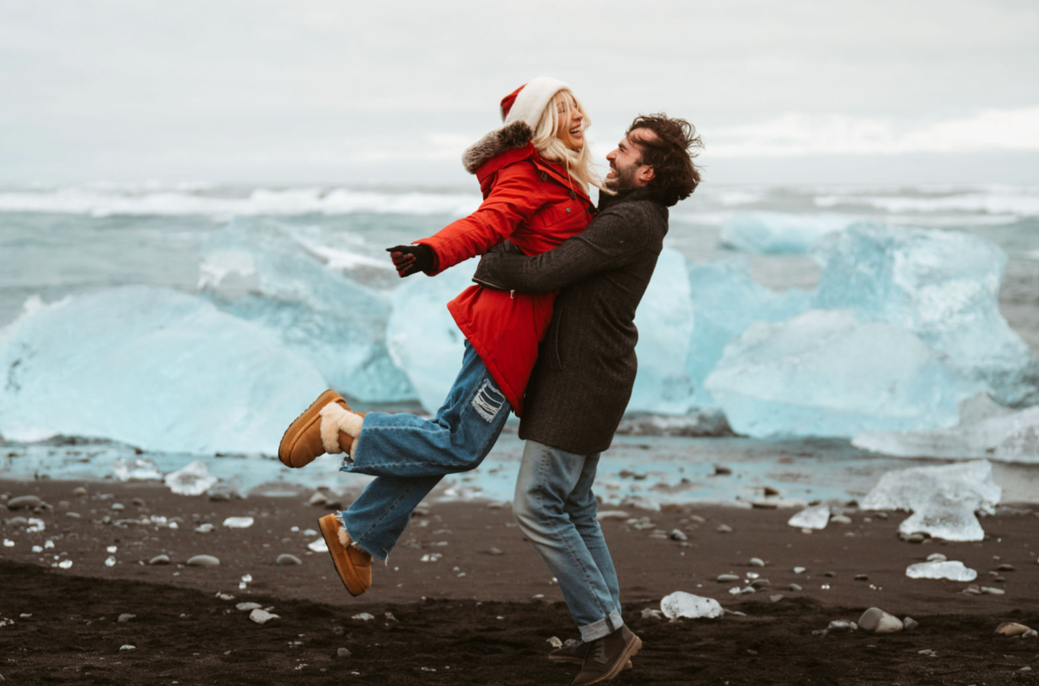 Couple embracing and laughing on black sand beach with turquoise glacier ice chunks in Iceland winter setting.