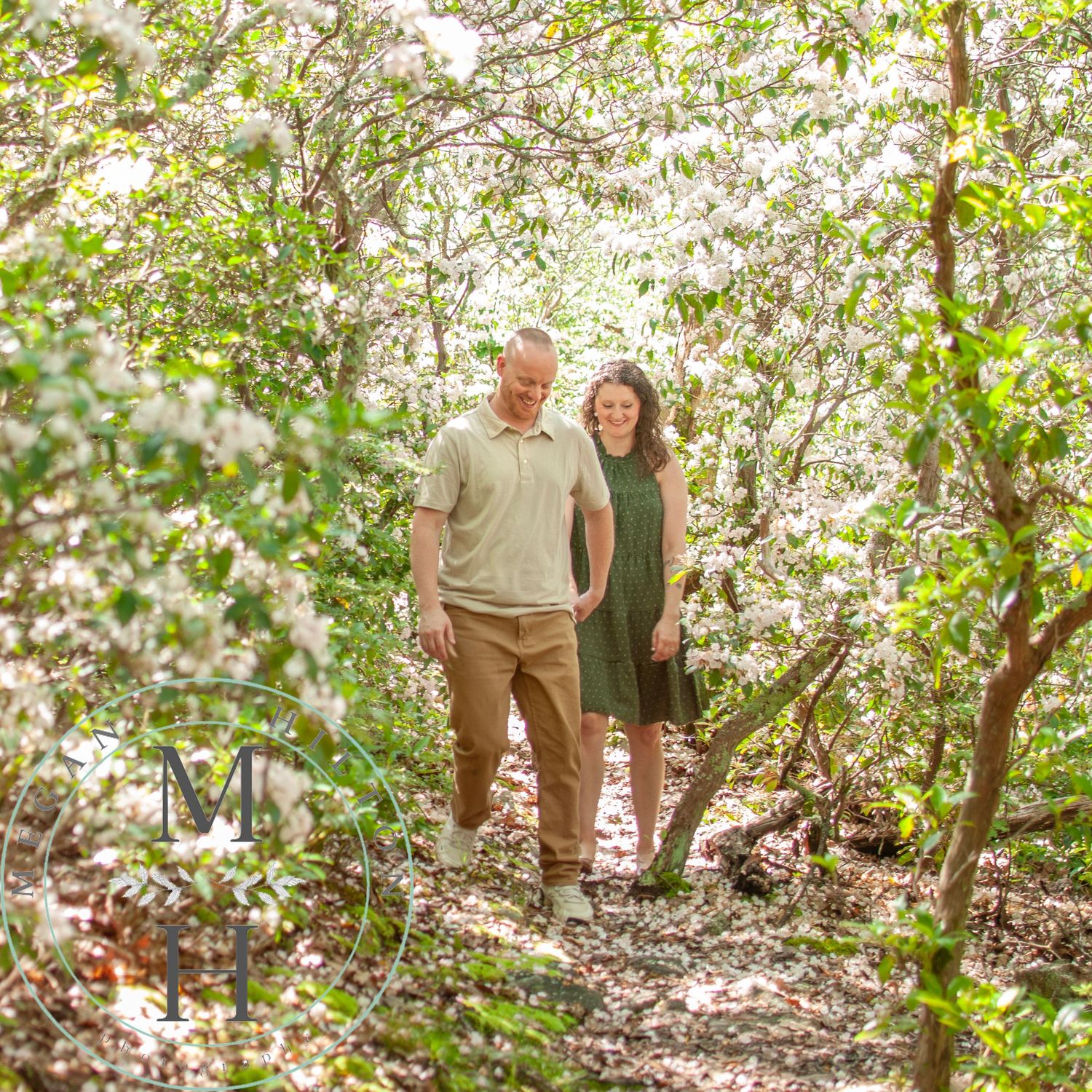 A couple walks together on a wooded trail surrounded by blooming white laurel trees.