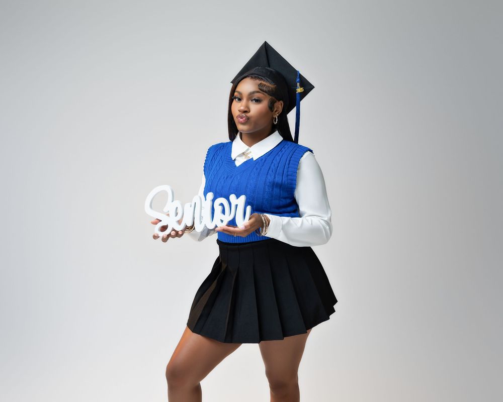 Graduate in blue sweater vest and black pleated skirt holds decorative 'Q/Moi' sign against gray backdrop.