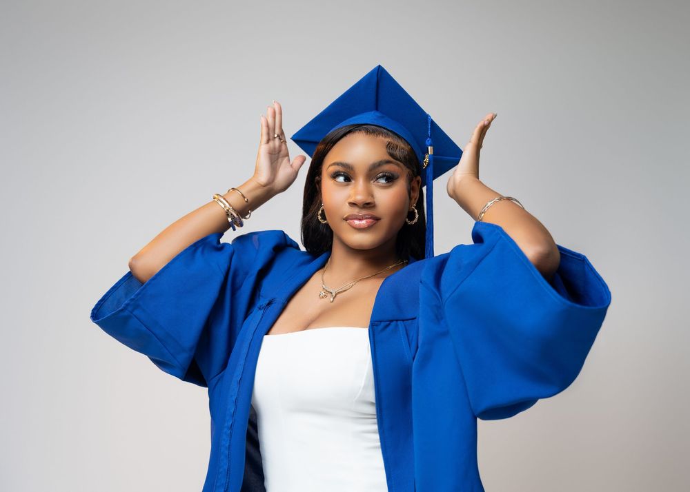 Someone in graduation attire poses with raised hands wearing a royal blue cap and gown with a white dress underneath.