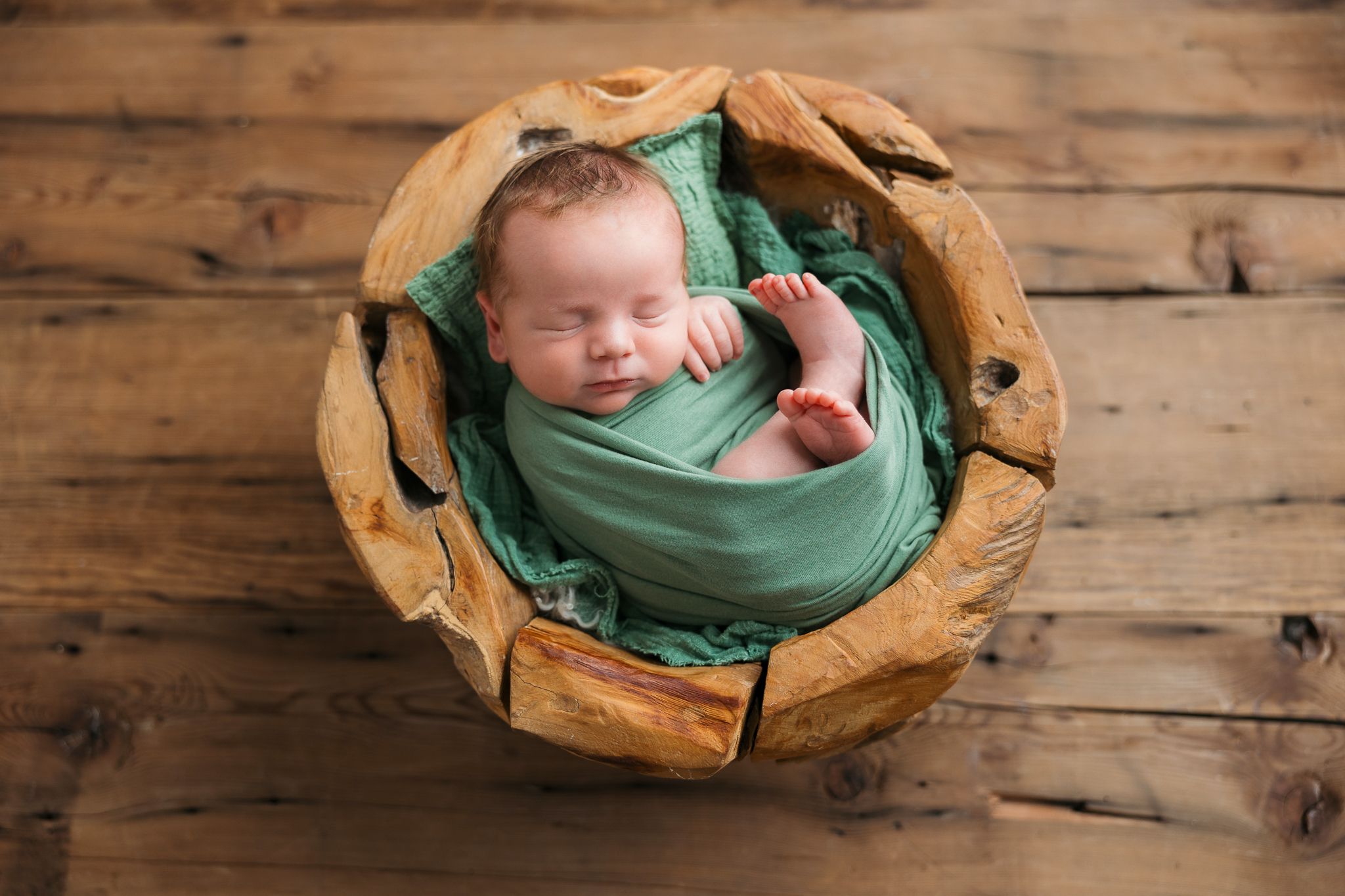 Newborn baby sleeps peacefully wrapped in green swaddle blanket inside wooden bowl on rustic floor.