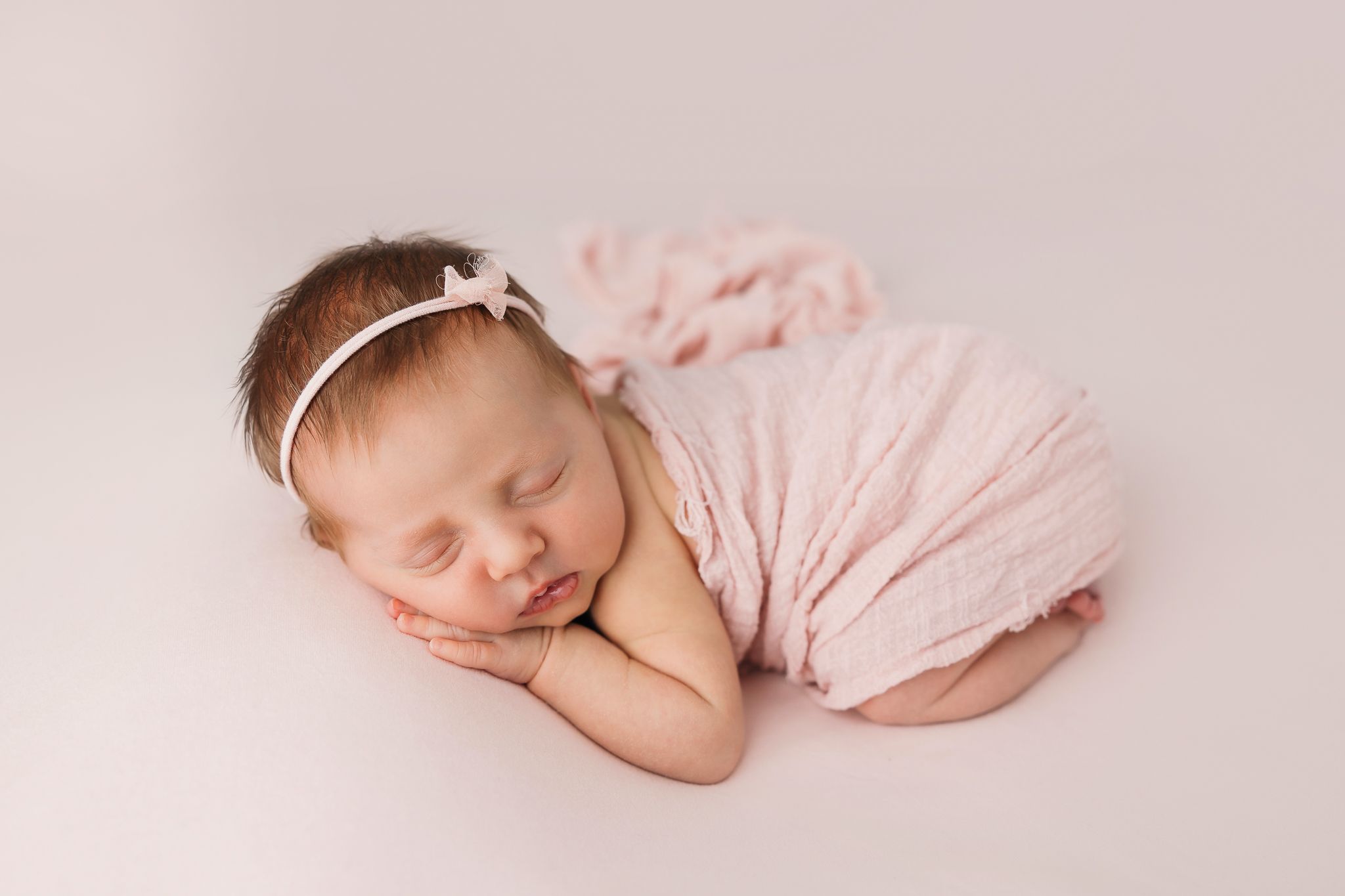 Sleeping newborn wrapped in soft pink fabric rests peacefully on light backdrop with delicate white headband.
