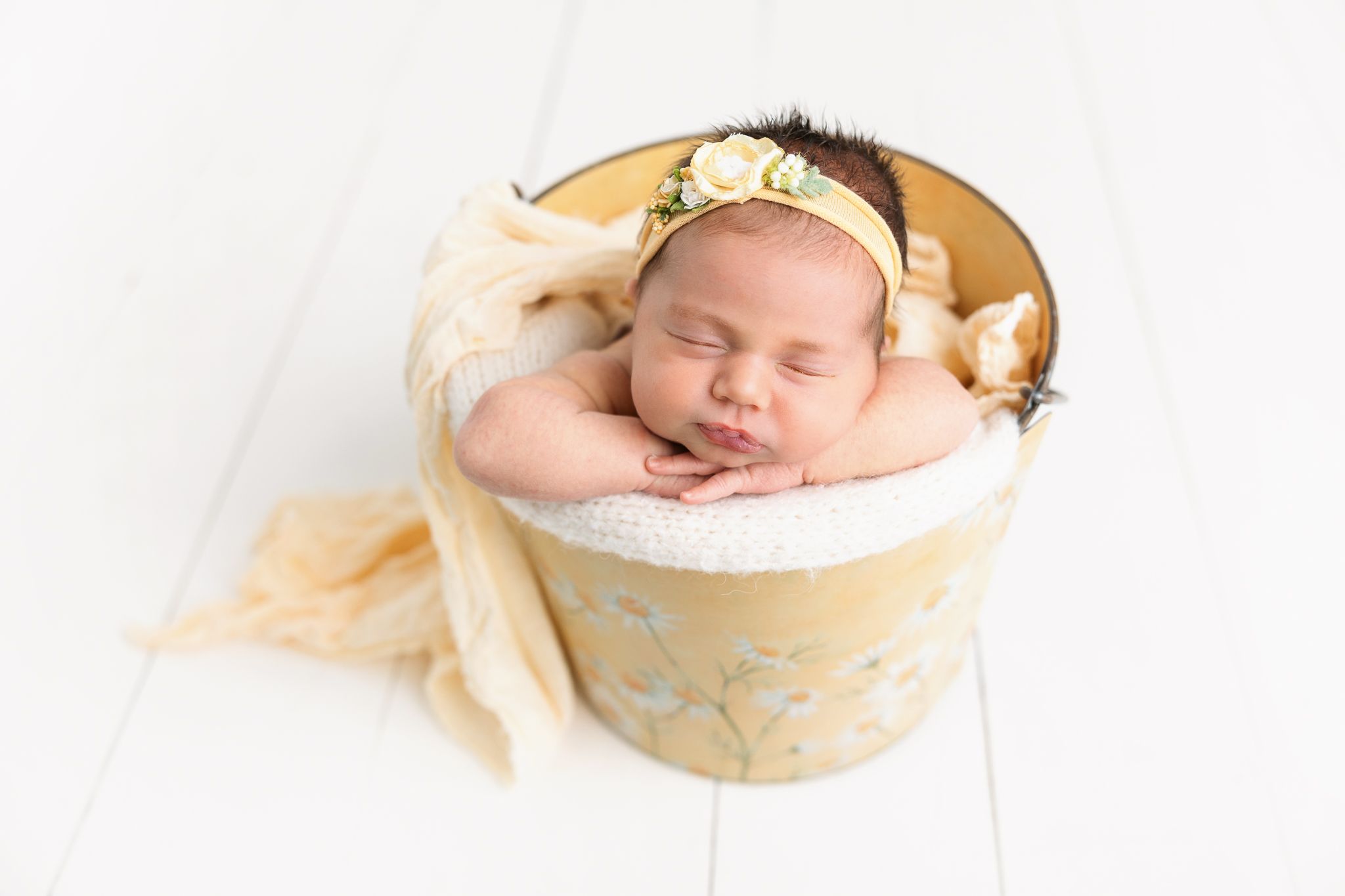 Peaceful sleeping newborn rests in vintage yellow container with soft fabric and floral details on white wooden background.