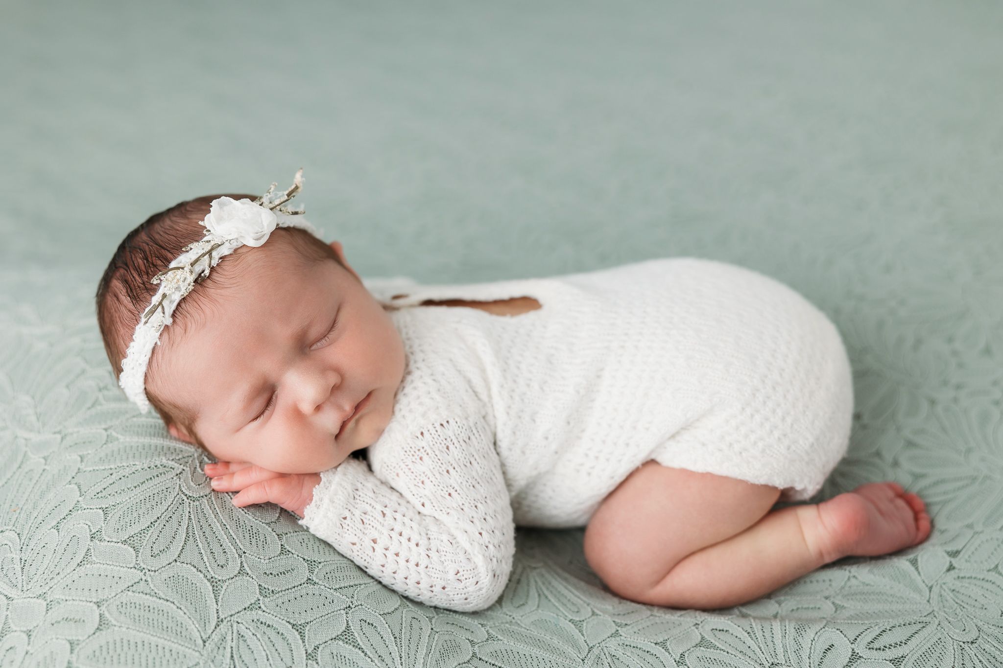Newborn baby wearing white lace outfit sleeps peacefully on grey blanket in professional portrait series.