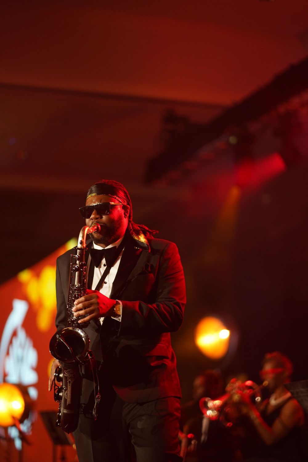 A musician plays saxophone on stage bathed in dramatic red lighting while performing at a concert.