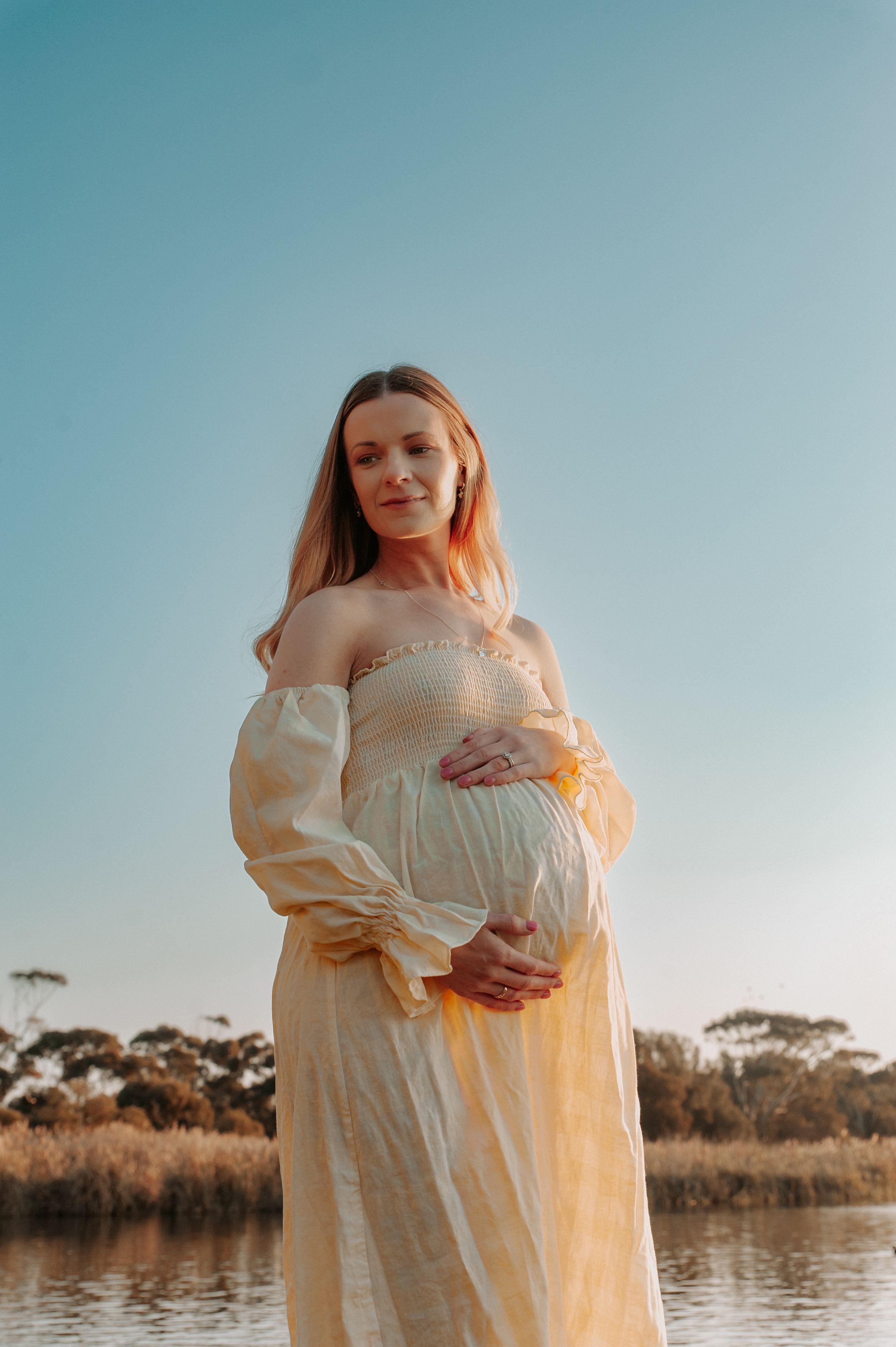 Pregnant woman in off-shoulder cream dress poses by waterside during golden hour.
Altona Family Photographer Melbourne
