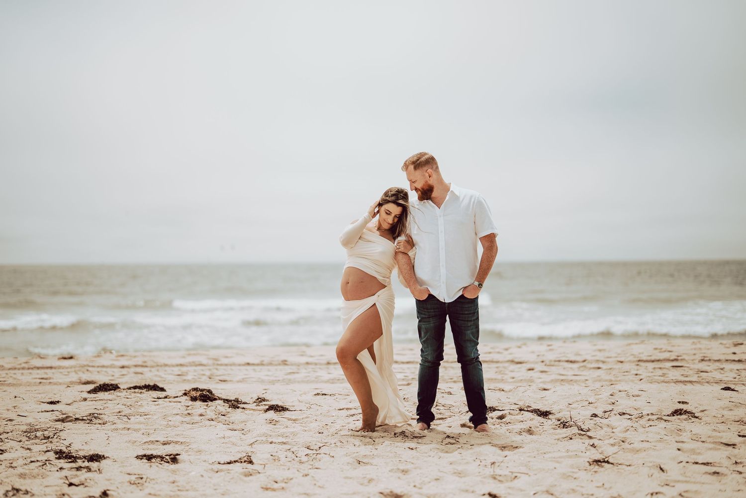 Pregnant couple posing on the sandy beach of Robert Moses, mommy in white maternity outfit, daddy in jeans and white shirt, ocean backdrop.