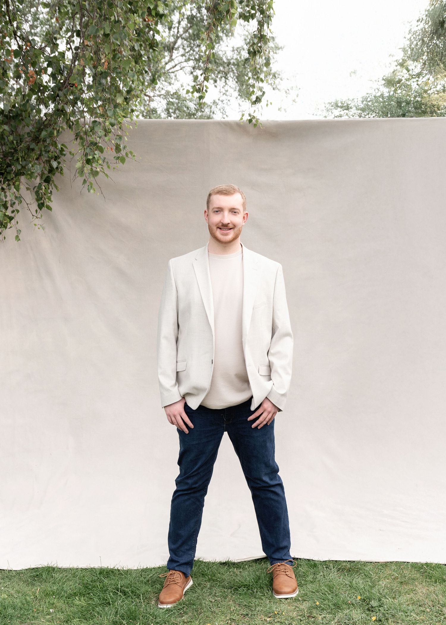 Man in white blazer wearing a mustache smiles and poses against light background.