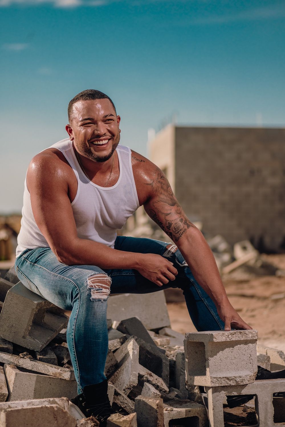 A person in a white tank top and ripped jeans sits smiling on concrete blocks against a bright blue sky.
