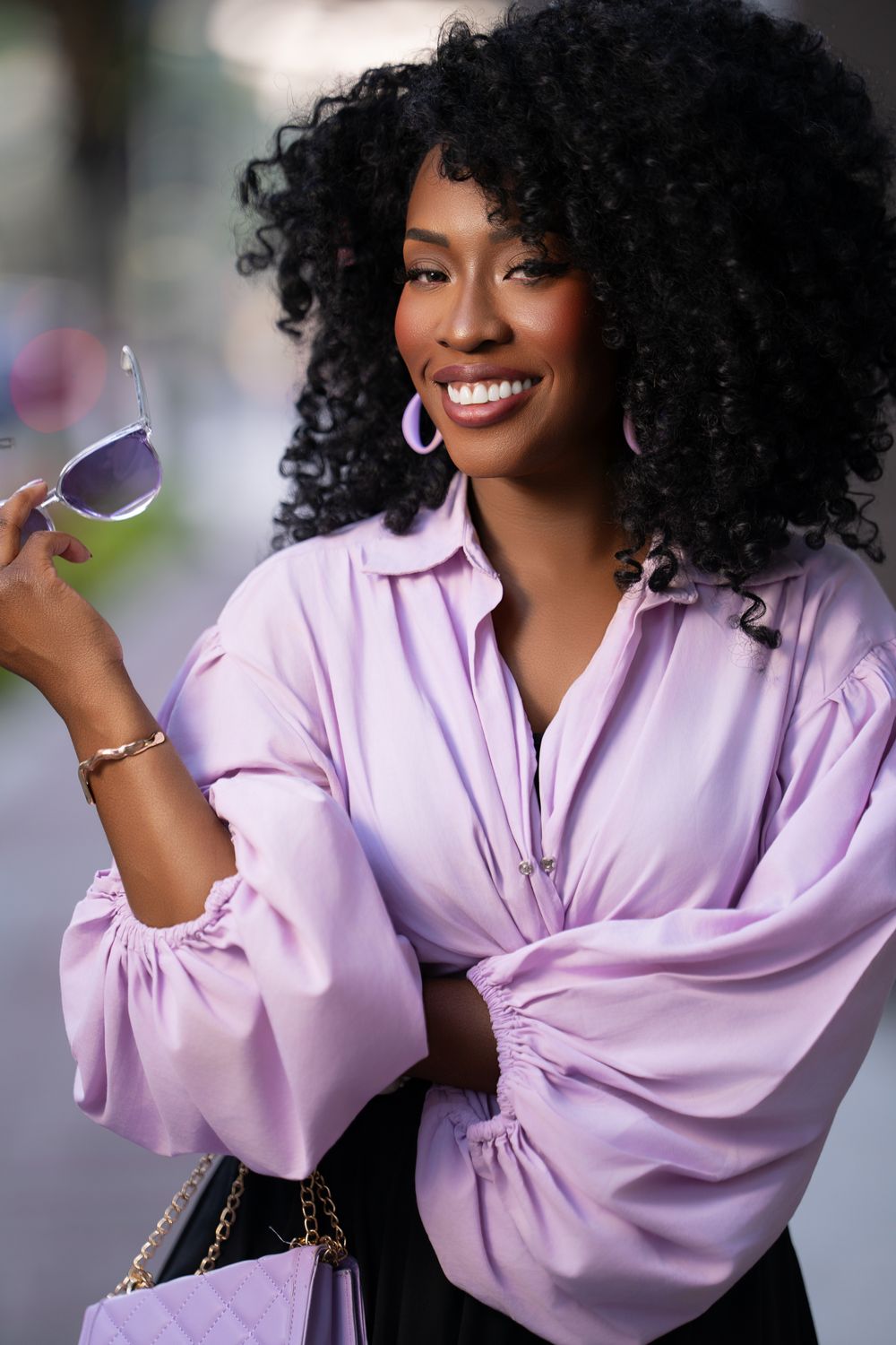 Fashionable woman in lavender blouse with voluminous curly hair and silver hoop earrings smiles while holding sunglasses.