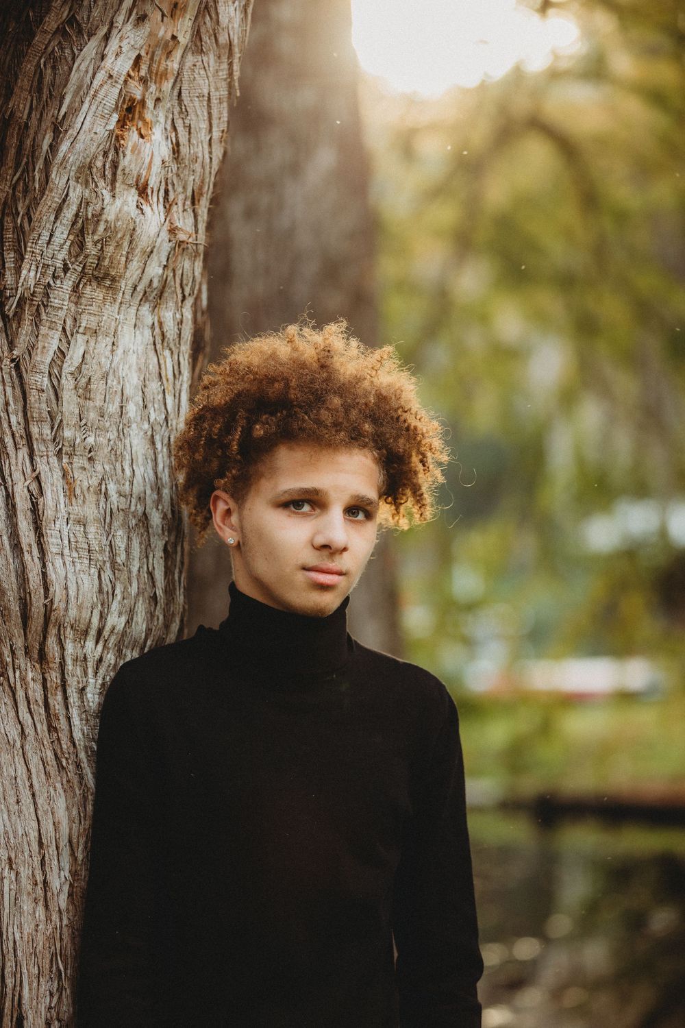 Portrait in natural lighting against a tree trunk wearing a black turtleneck with curly hair in autumn setting.
