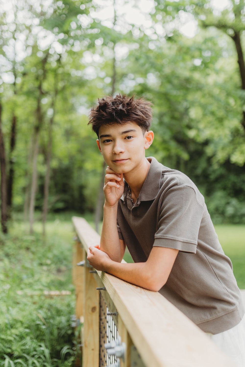 Young person in a gray polo shirt leaning thoughtfully on a wooden railing in a lush green outdoor setting.