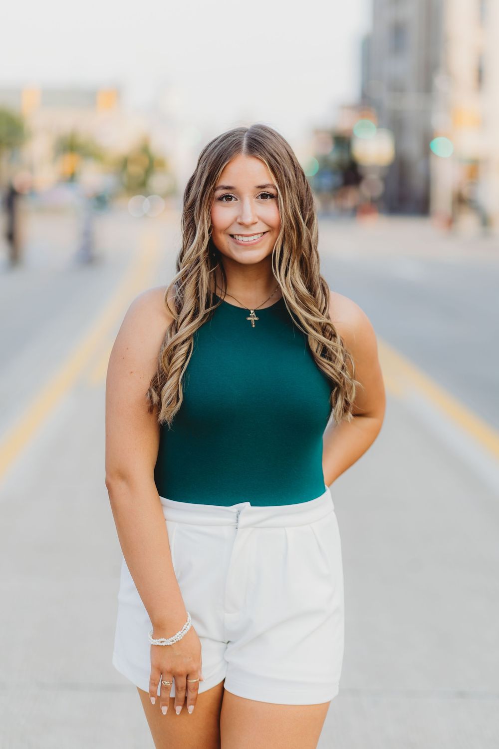 A person stands on a city street wearing a green sleeveless top and white shorts with long wavy hair.