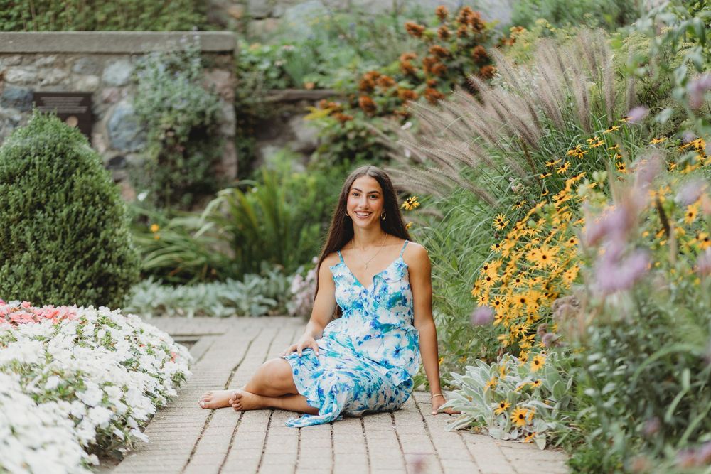 Person in blue floral dress sits on wooden pathway surrounded by vibrant summer garden flowers.
