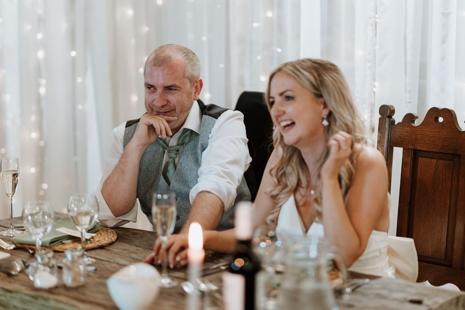 Wedding reception guests seated at a candlelit table with twinkle lights in the background.