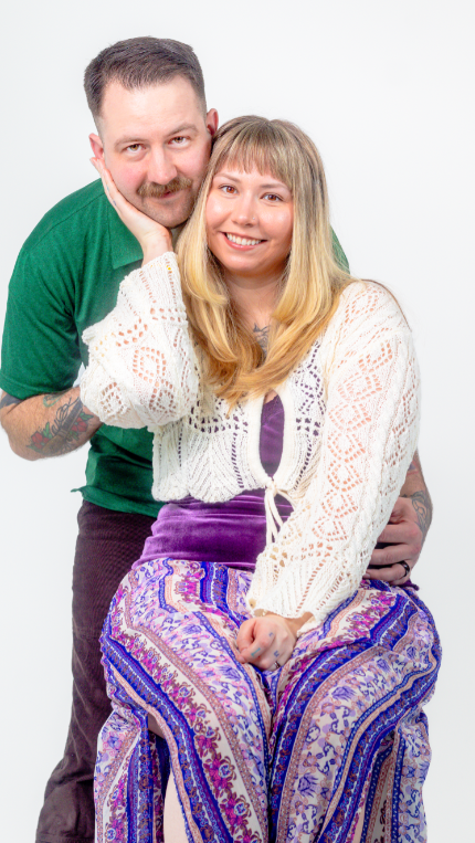 Couple posing together against white background wearing green sweater and white lace top with purple patterned pants.