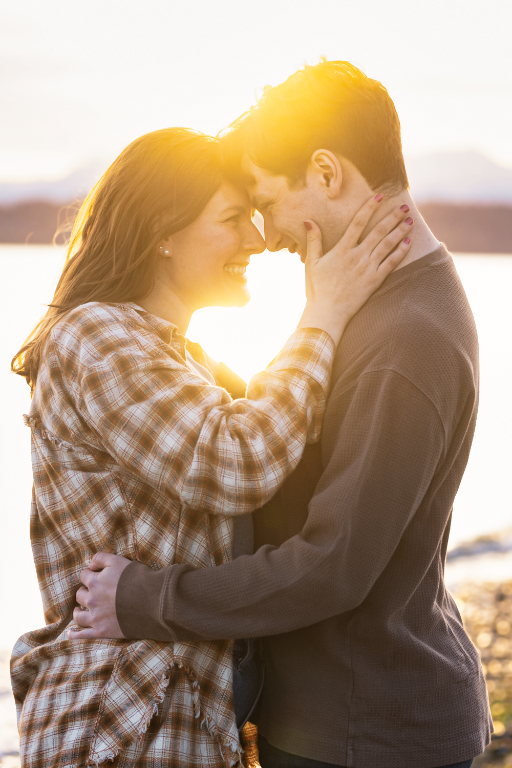 Couple sharing an intimate moment at sunset on a beach, silhouetted against golden sunlight through a plaid shirt.