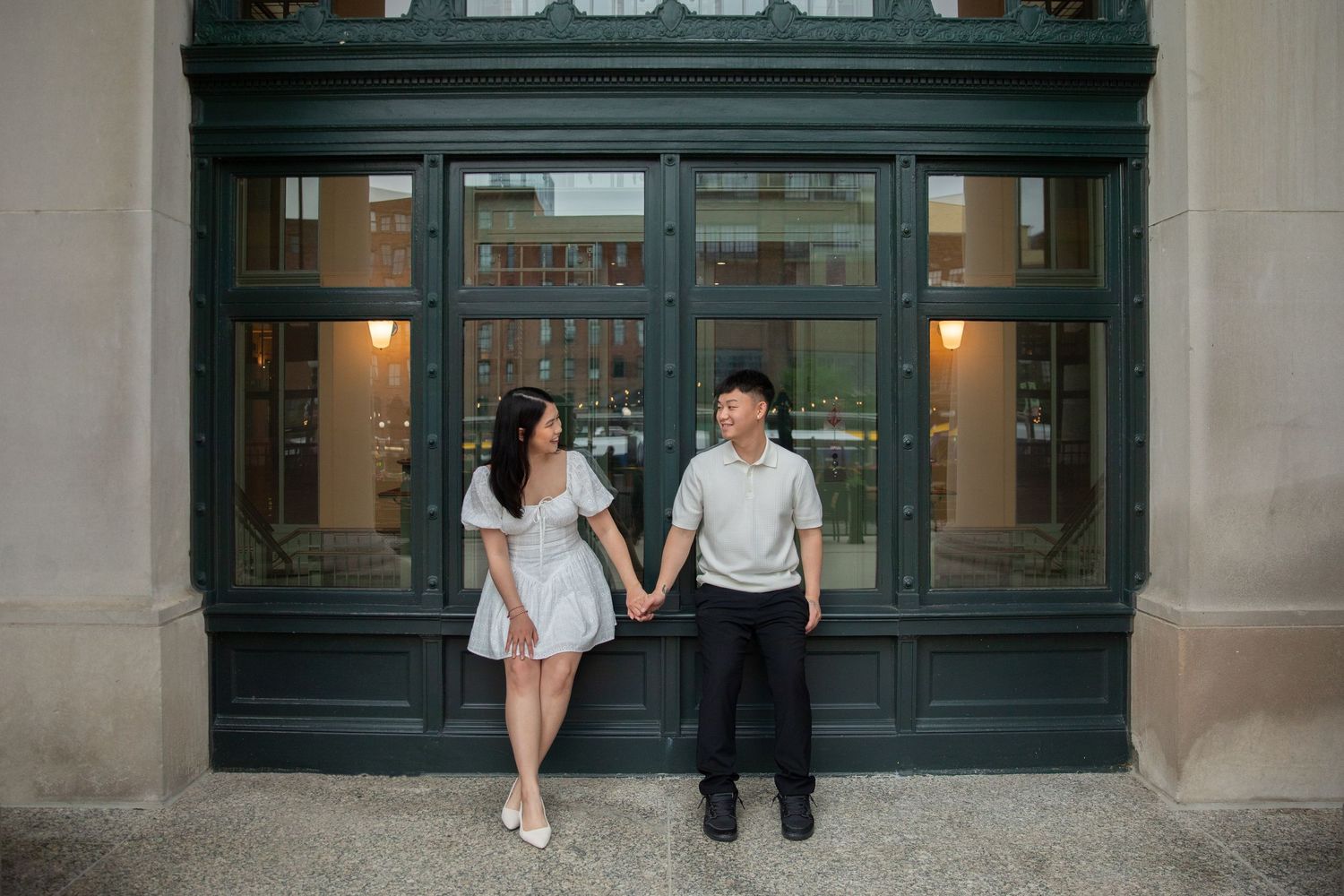 A couple holds hands while standing in front of a classic dark green storefront entrance with glass paneling.