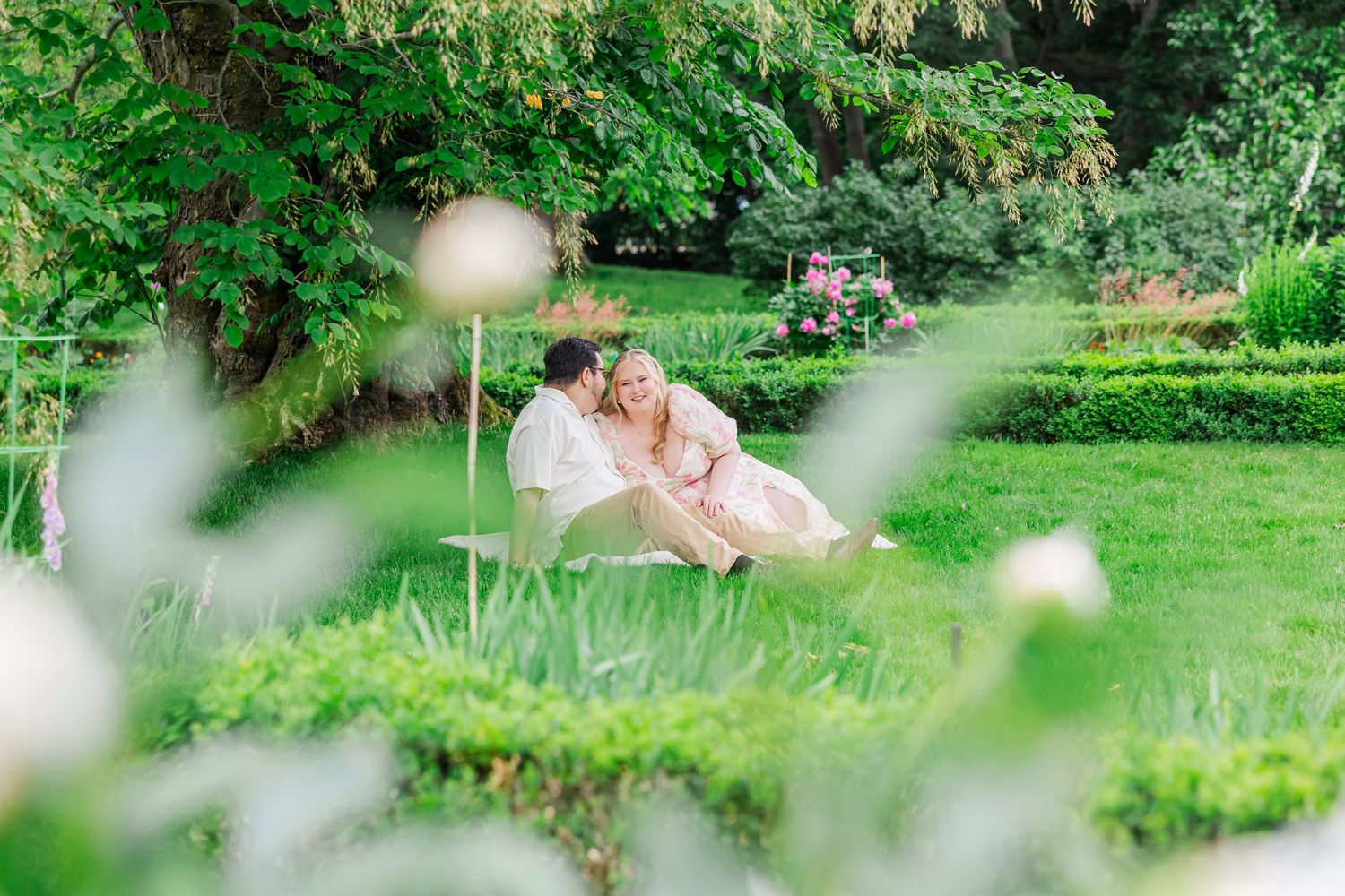 Two people in pastel attire share a romantic moment on lush green garden lawn framed by blurred flowers and foliage.