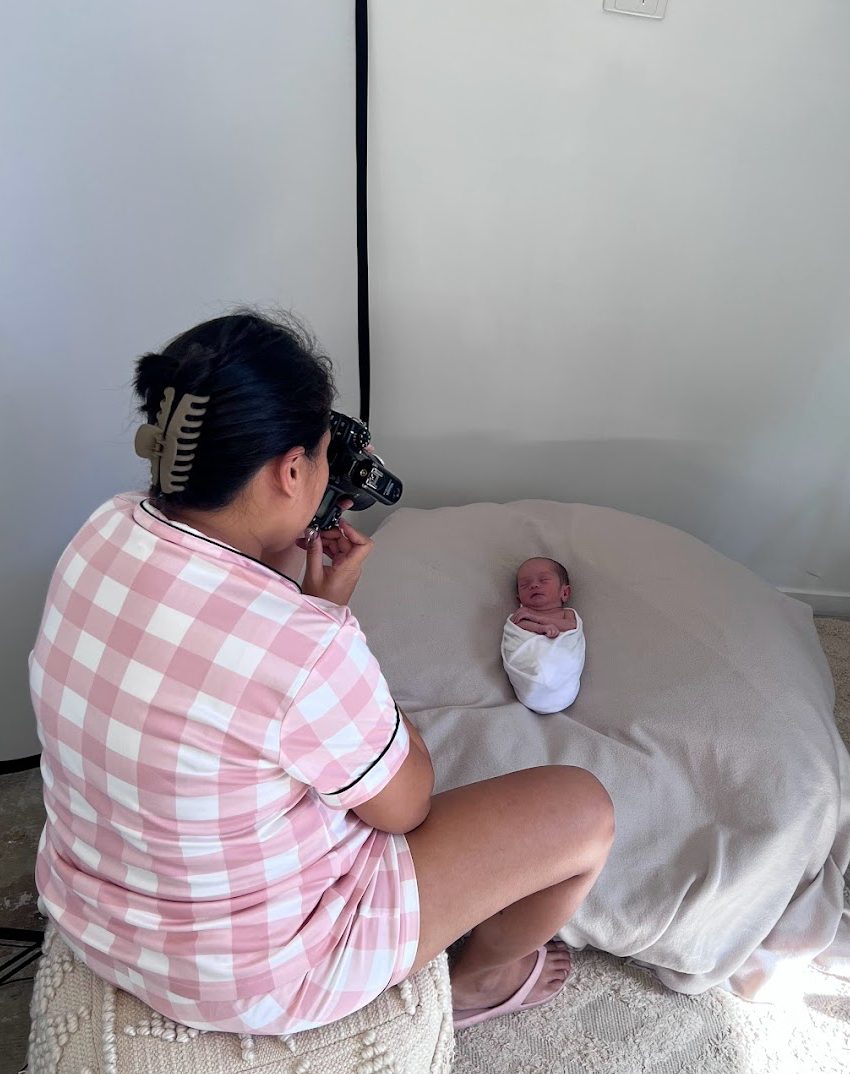 Professional photographer wearing a pink gingham shirt takes photos of swaddled newborn on grey bedding.