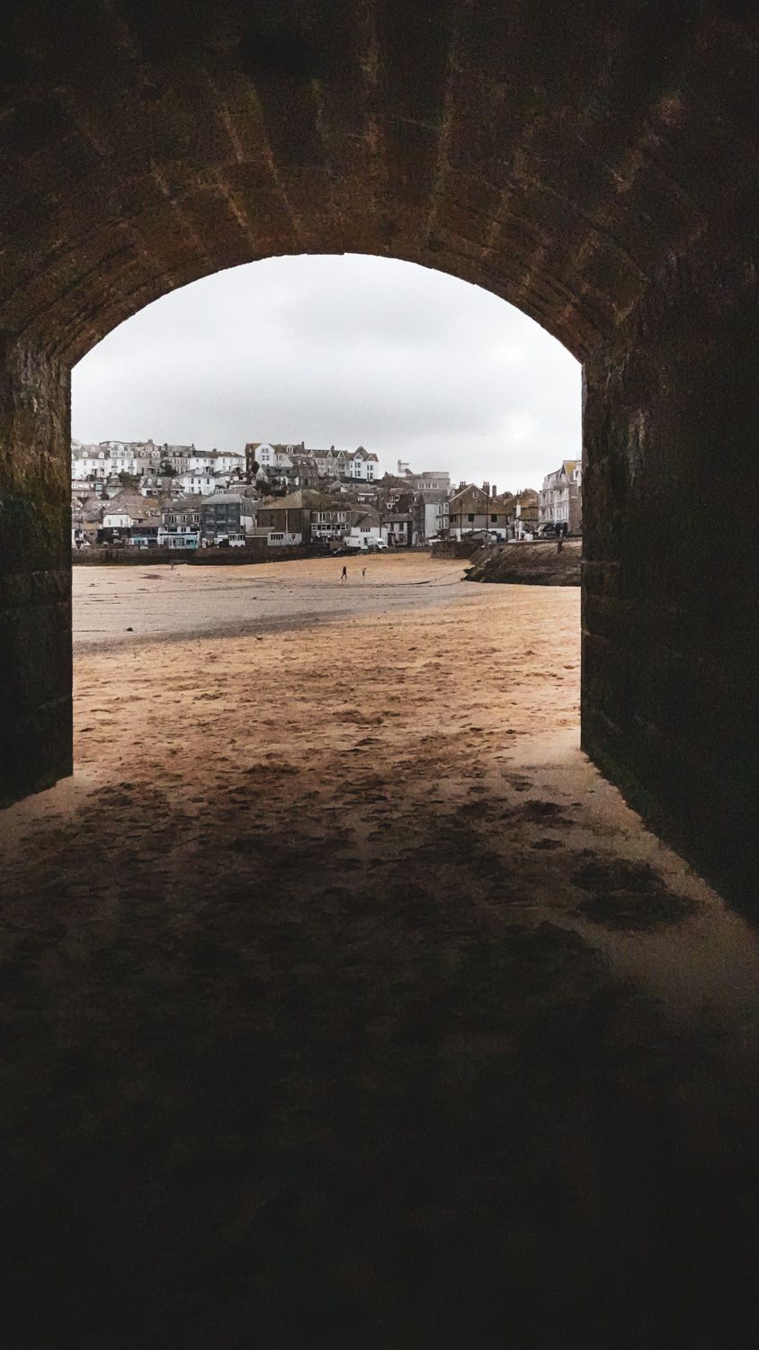 Panoramic view of a sandy beach from inside a stone archway tunnel with coastal buildings visible in the background.