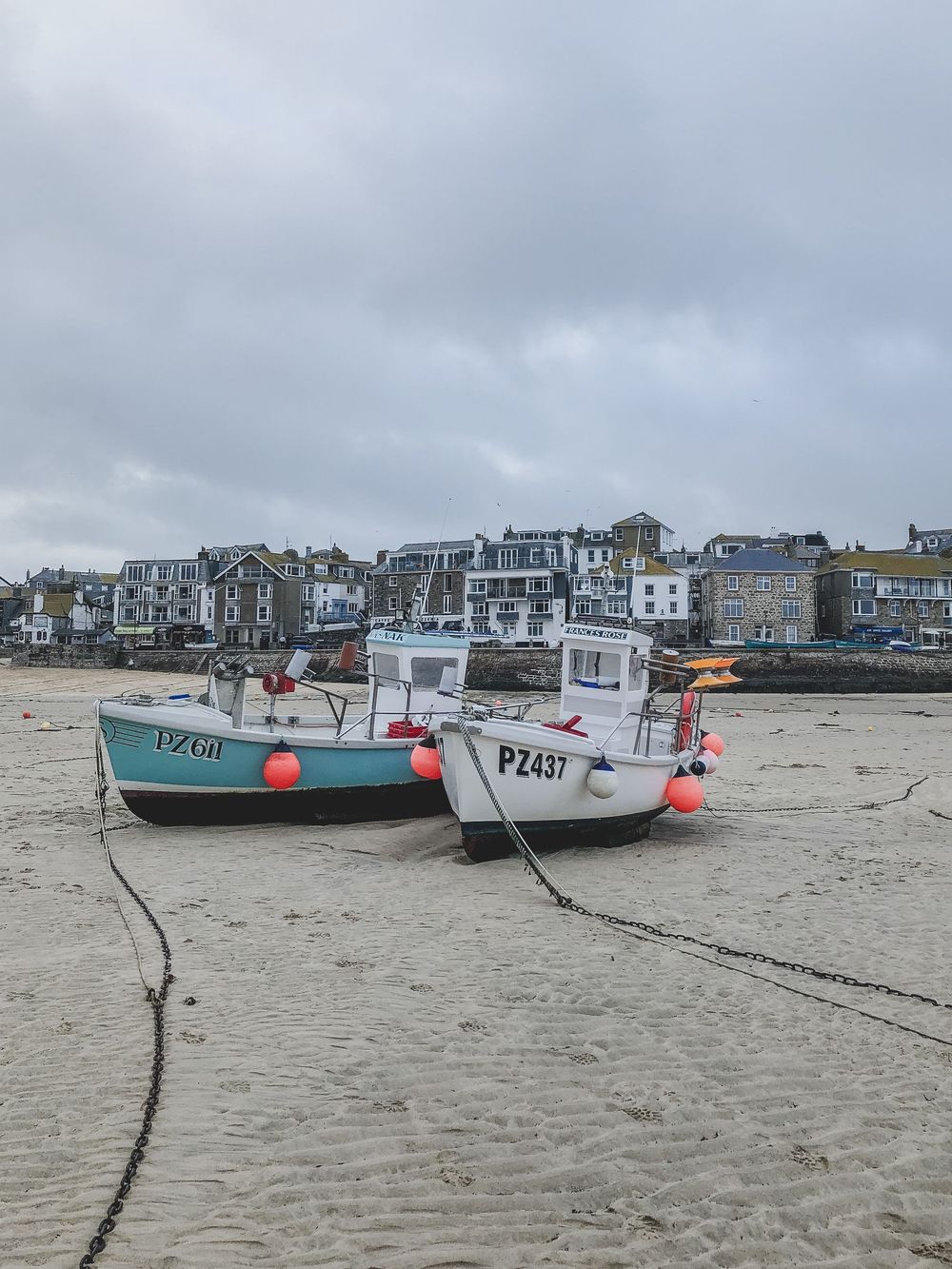 Small fishing boats rest on sandy beach with coastal houses and cloudy sky in the background.