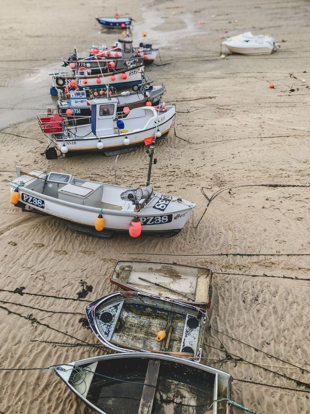 Small fishing boats with buoys and flags line up at low tide on a sandy beach shore.