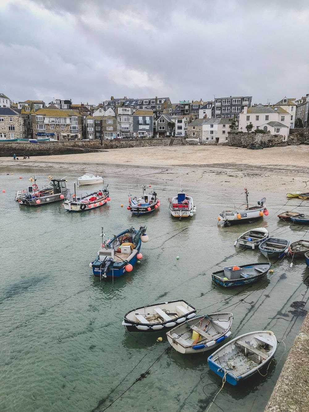 Small fishing boats moored in a harbor during low tide with coastal buildings and cloudy skies in Cornwall.