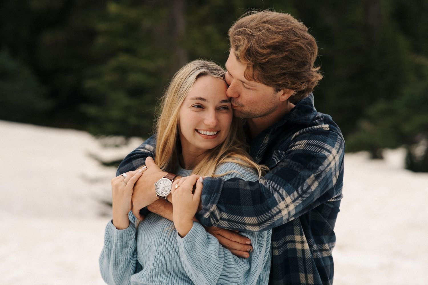 A couple embraces and laughs in the snow while wearing winter clothing during a romantic outdoor photo session.